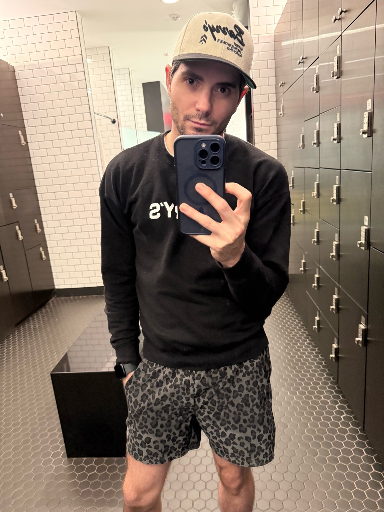 A man takes a selfie in a locker room, wearing a baseball cap, sweatshirt, and leopard print shorts. He appears tired.