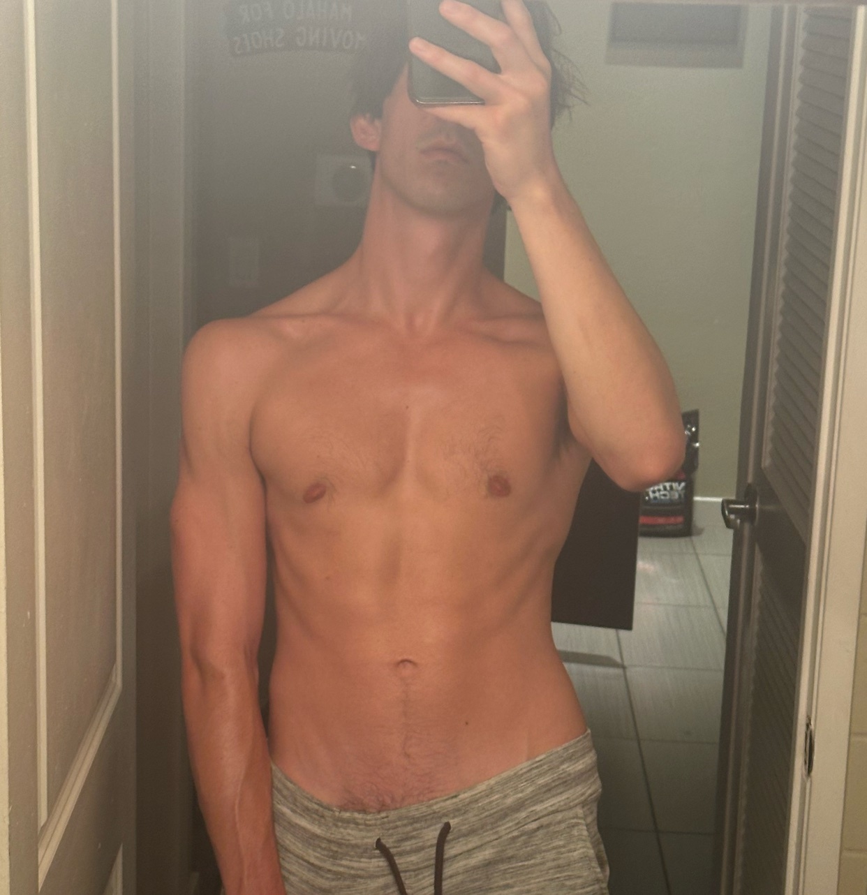 A man takes a shirtless mirror selfie, showcasing his physique. He is wearing grey sweatpants.