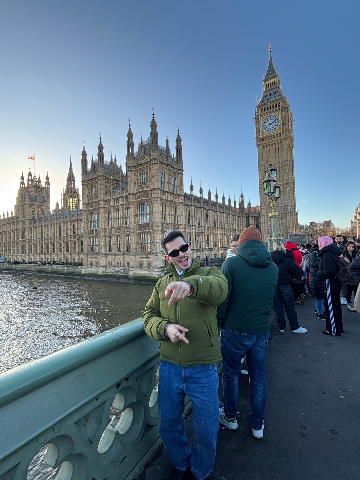 A man poses for a photo in front of the Houses of Parliament and Big Ben in London. The sun is setting, casting a warm glow on the scene.