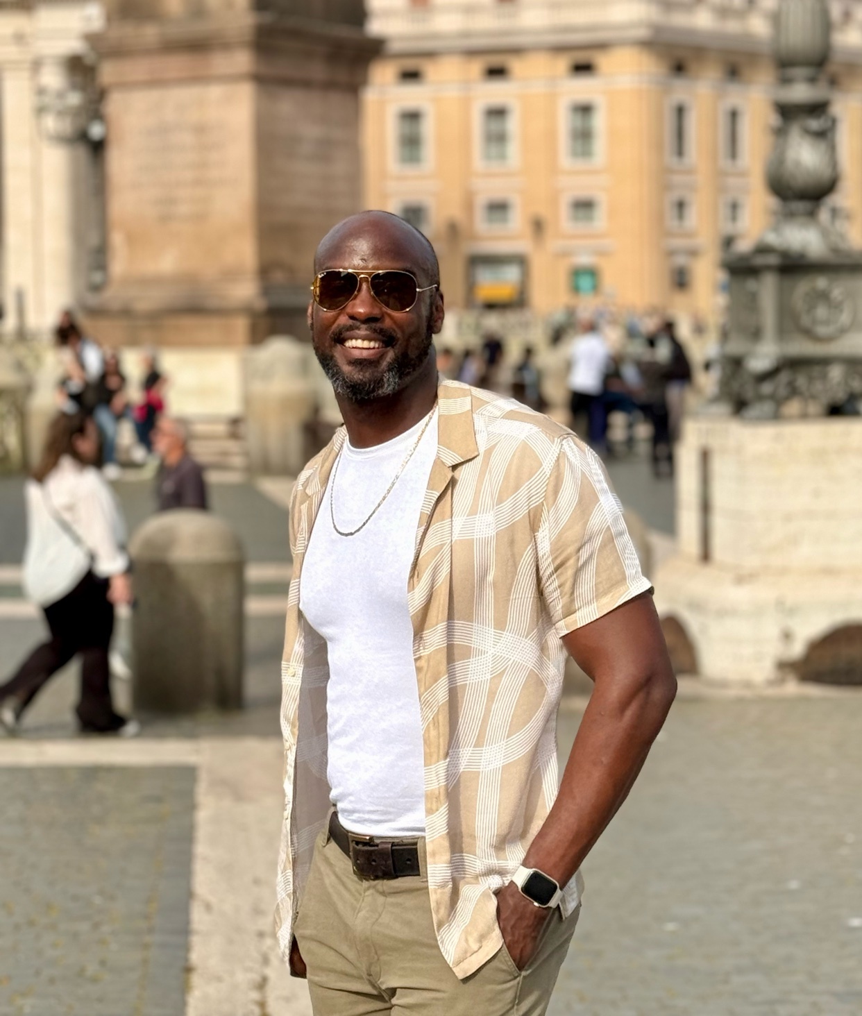 A man poses for a photo in front of a building. He is wearing sunglasses and a stylish outfit.