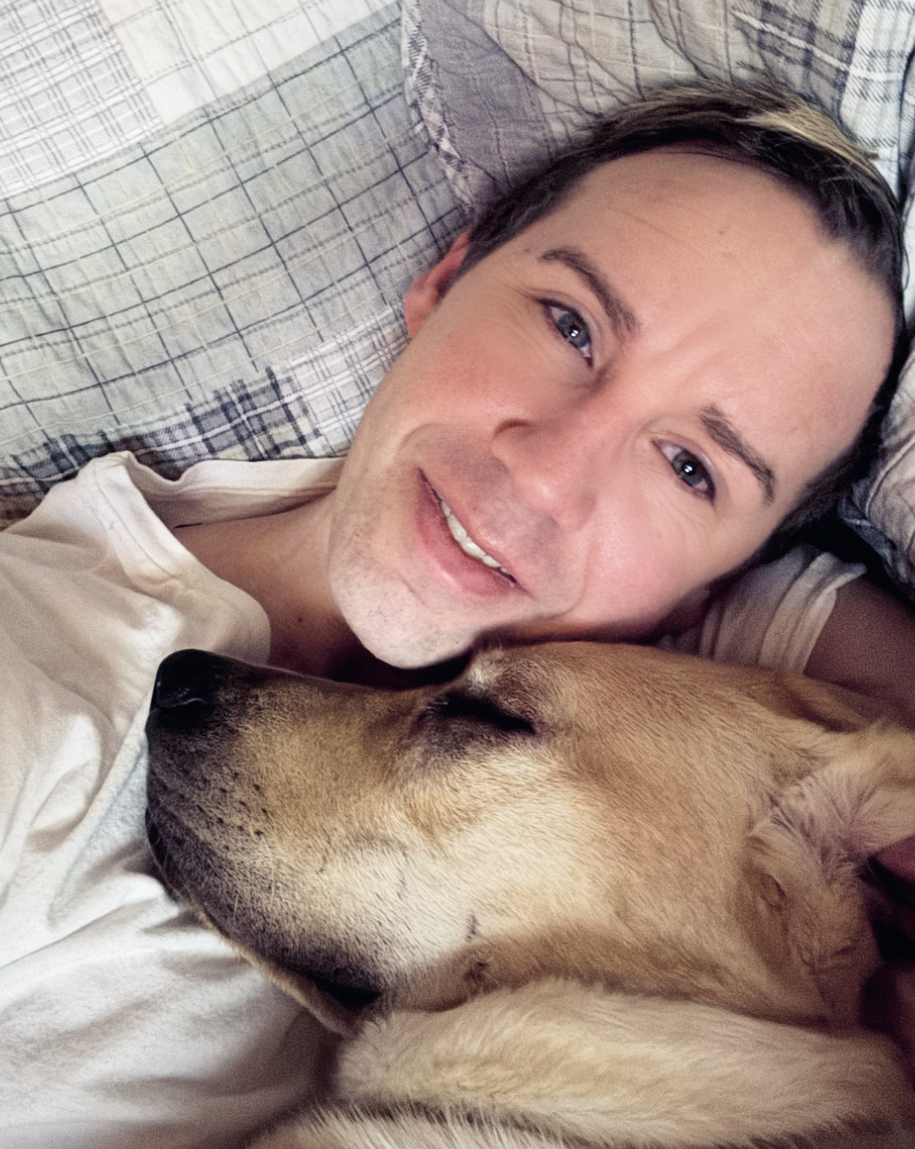 A man and his dog, Watson, are cuddling in bed. The man is smiling at the camera while his dog rests its head on his chest.