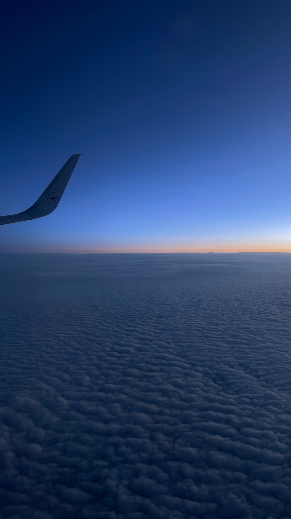An aerial view of the sky and clouds from an airplane window. The wing of the plane is visible in the upper left corner.