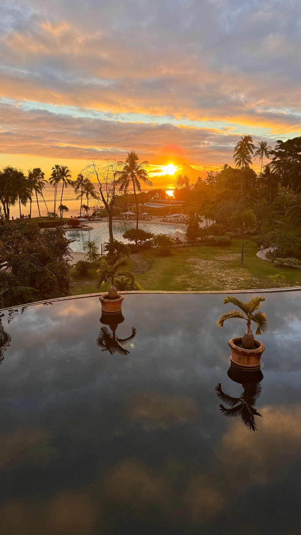 A vibrant sunset paints the sky with orange and yellow hues over a tropical resort. Palm trees line the coast, and their reflections are mirrored in the still water of an infinity pool in the foreground.