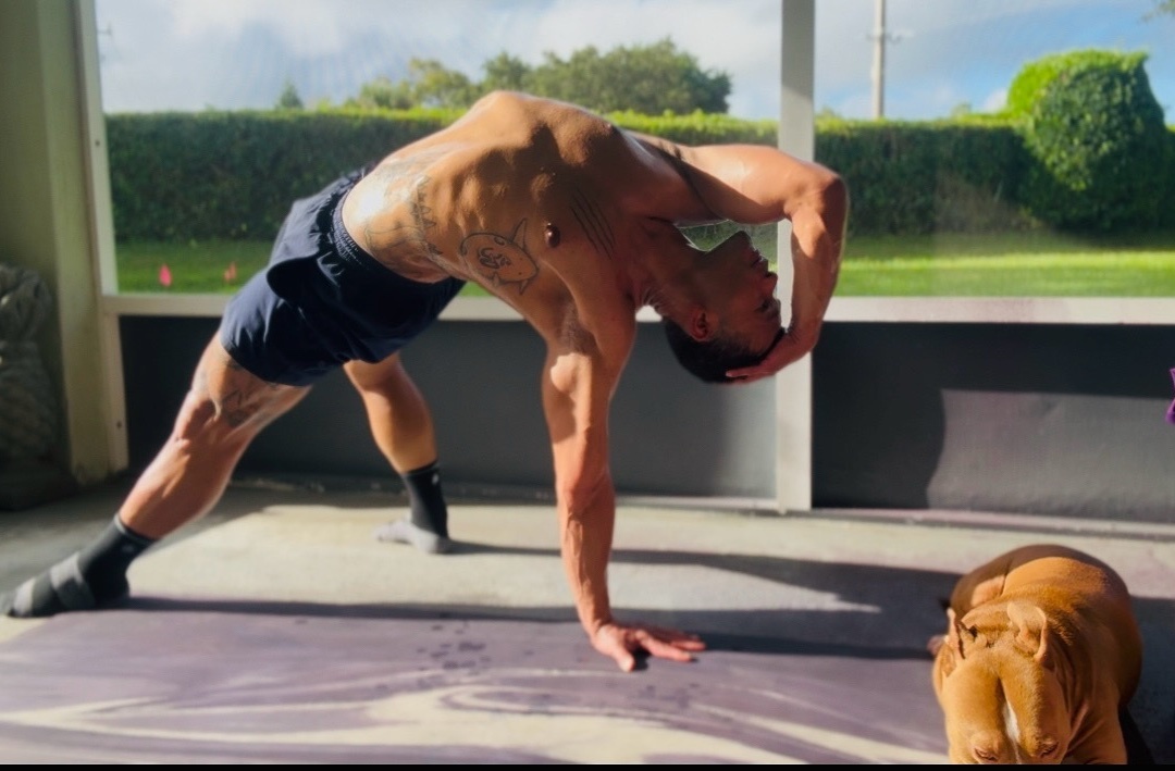 A man is doing a yoga pose in a gym. He is shirtless and muscular, with a tattoo on his back.
