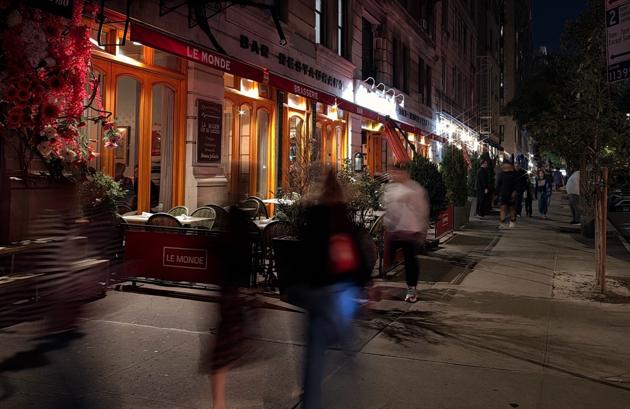 A blurred motion shot of people walking past a restaurant and bar called Le Monde at night. The restaurant has warm lighting and outdoor seating.
