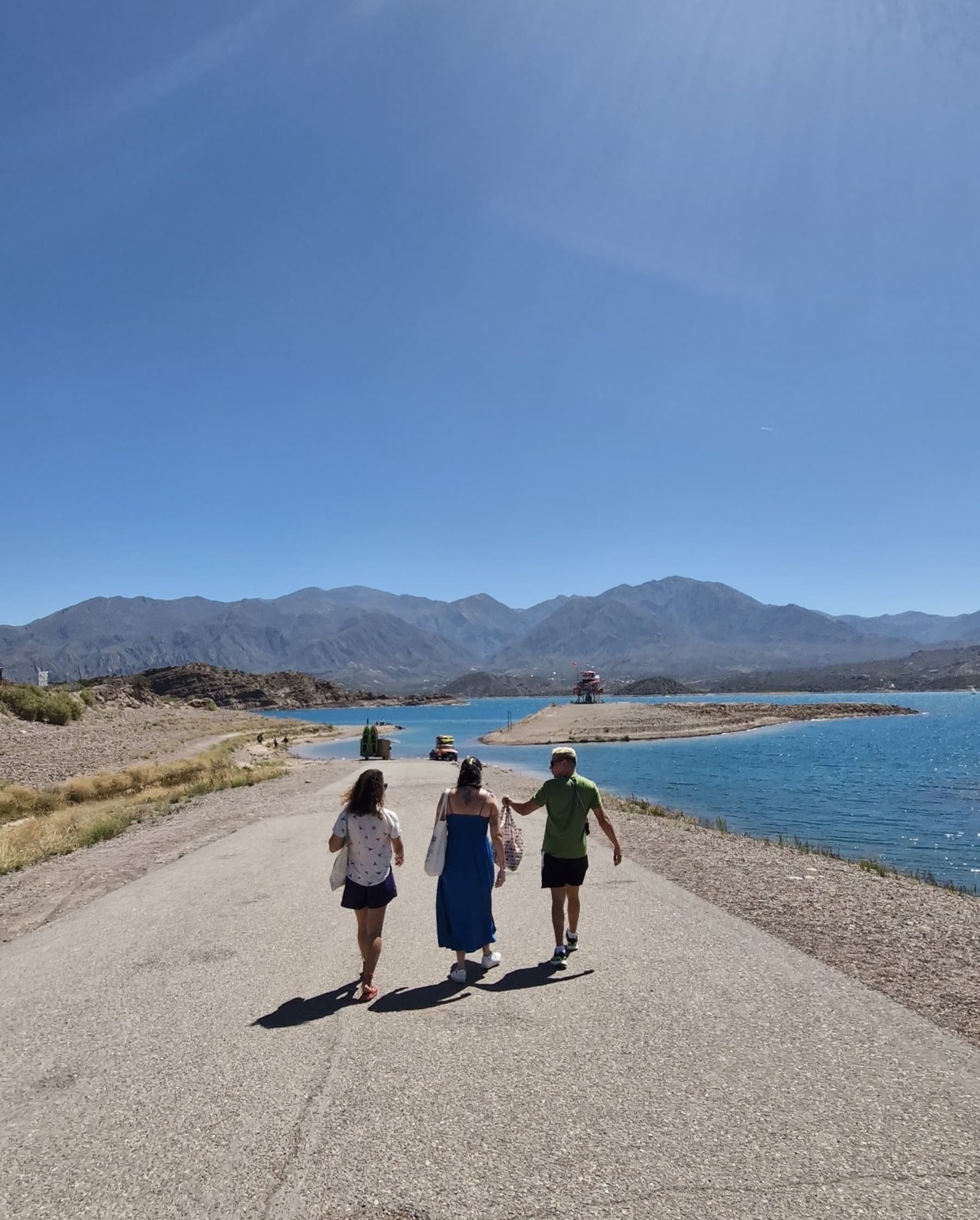 Three people walk away from the camera down a paved path next to a bright blue lake. Mountains are visible in the distance under a clear, sunny sky.