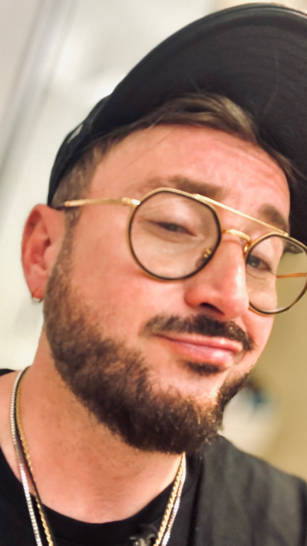 A close-up selfie of a man wearing round glasses, a black baseball cap, and gold chains around his neck. He has a beard and stubble, and is looking slightly to the side with a playful expression.