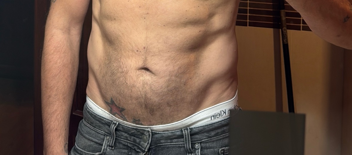 A close-up shot focuses on a man's torso, revealing his abs and a tattoo on his hip. He is wearing jeans and white Calvin Klein underwear.