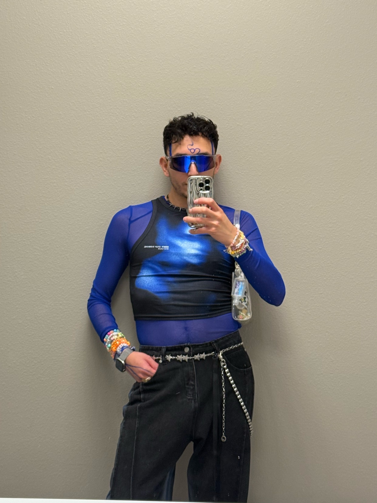A man takes a mirror selfie, showcasing his outfit. He wears a blue long-sleeved shirt, sunglasses, and jewelry.