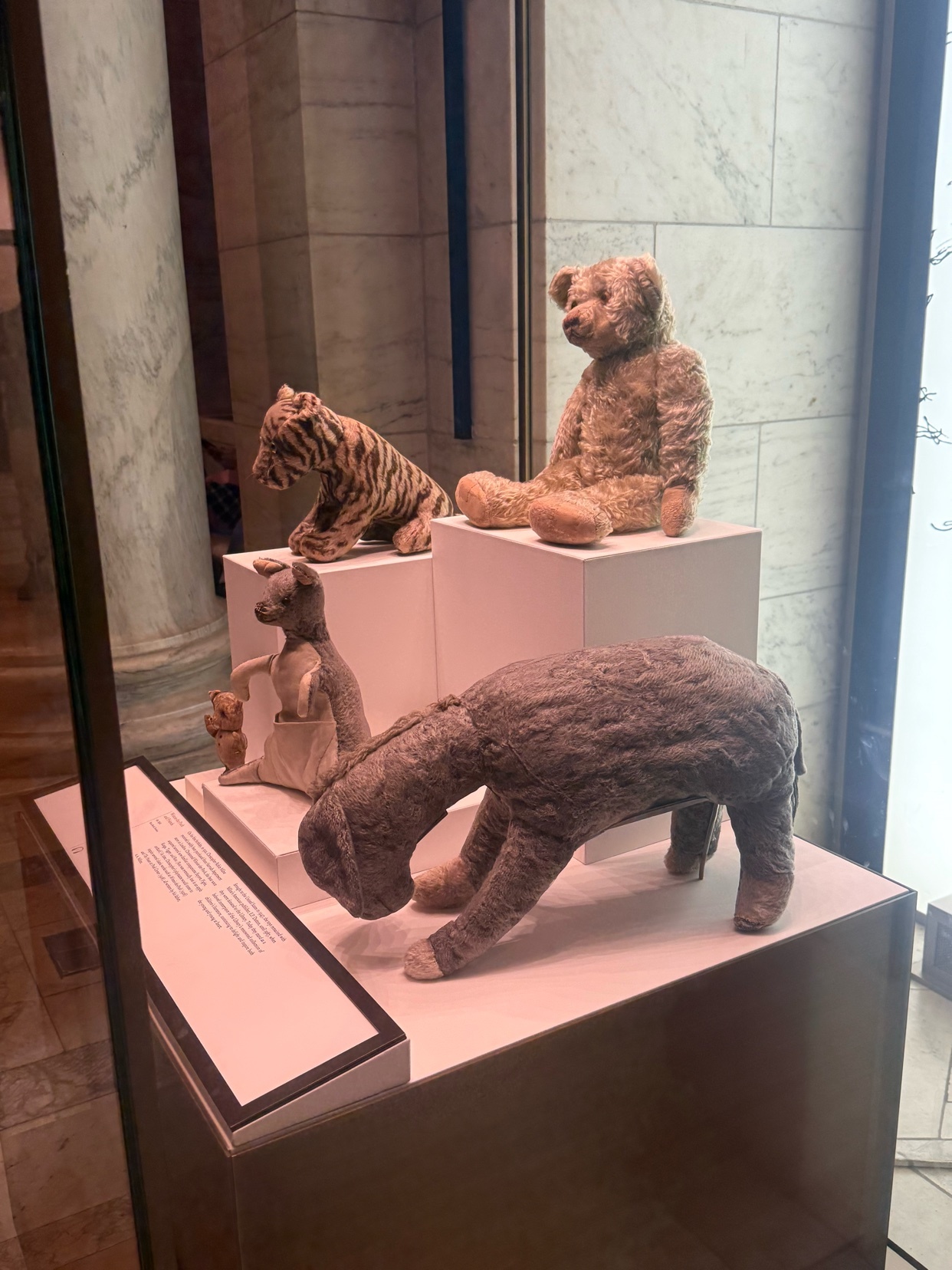 A museum display showcases several beloved Winnie the Pooh characters, including Eeyore, Tigger, Kanga, Roo, and a classic teddy bear, all presented as stuffed animals on white pedestals.