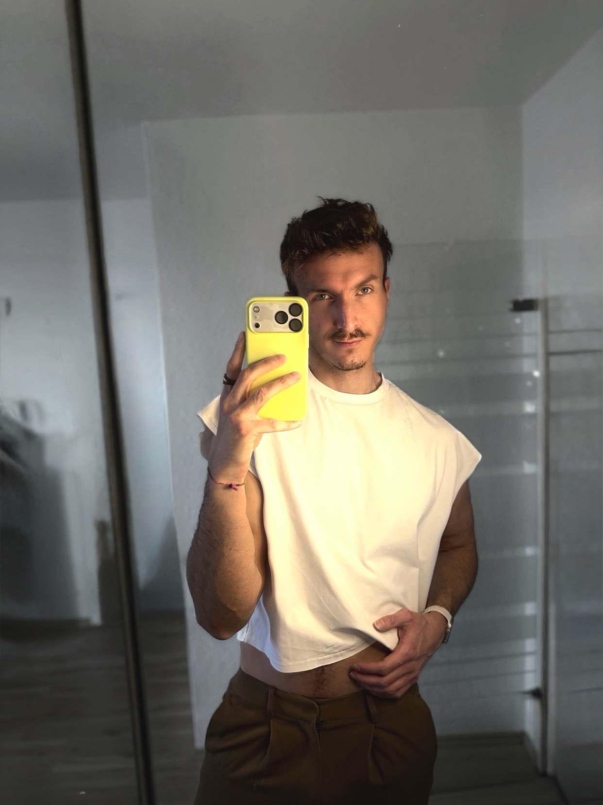 A man takes a mirror selfie, showing off his physique. He is shirtless, wearing brown pants and a white t-shirt.