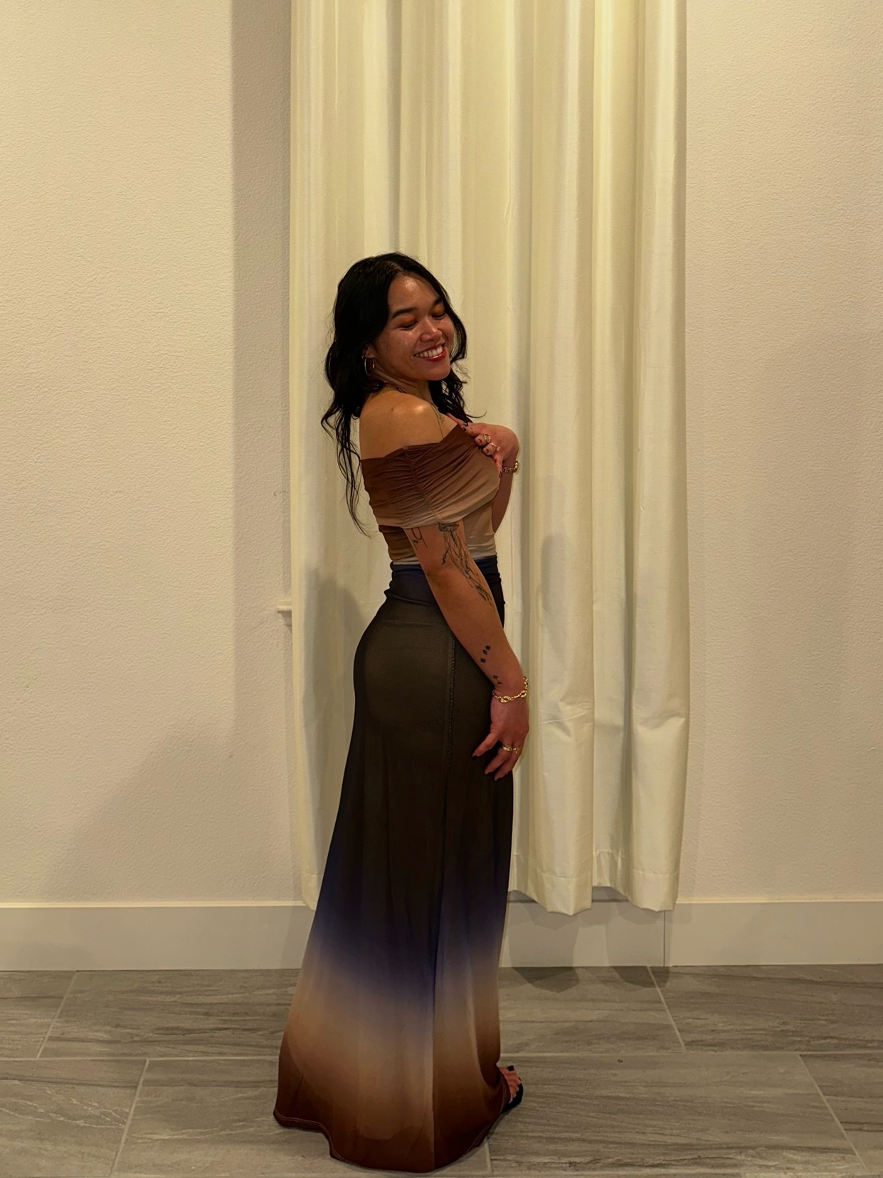 A woman poses in a formal dress, smiling at the camera. The dress has an ombre effect, and she is standing in front of a curtain.