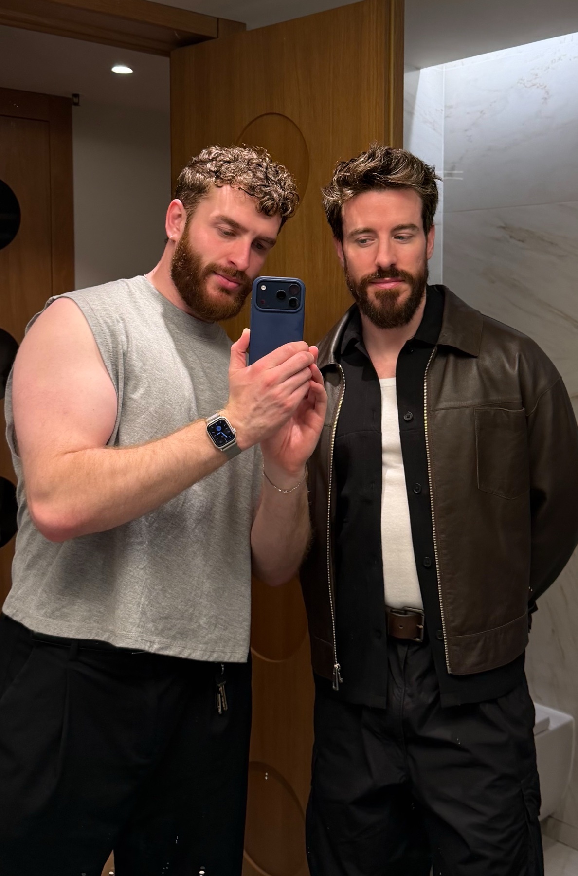 Two men pose for a selfie in a modern bathroom. The man on the left wears a grey sleeveless shirt and black pants, while the man on the right wears a brown leather jacket over a black shirt and pants.