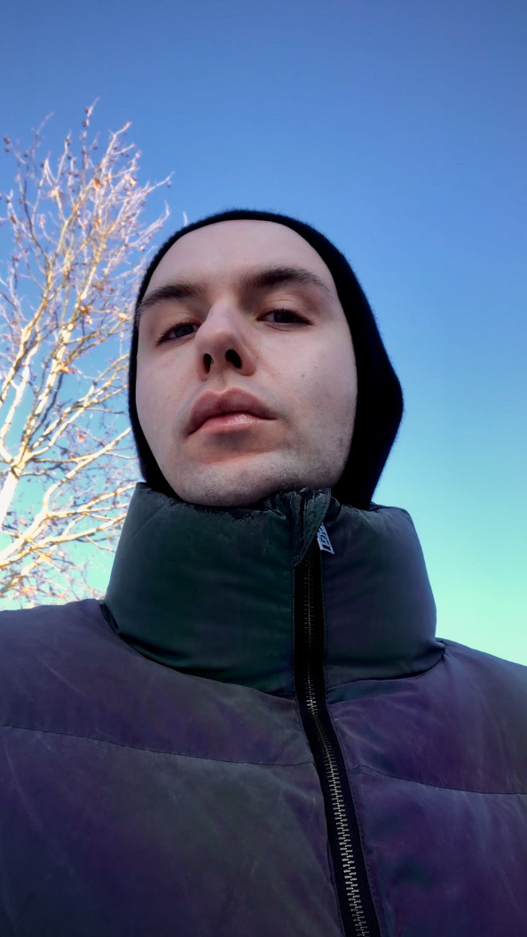 A man takes a selfie outdoors on a cold winter day. He is wearing a black hoodie and a jacket.