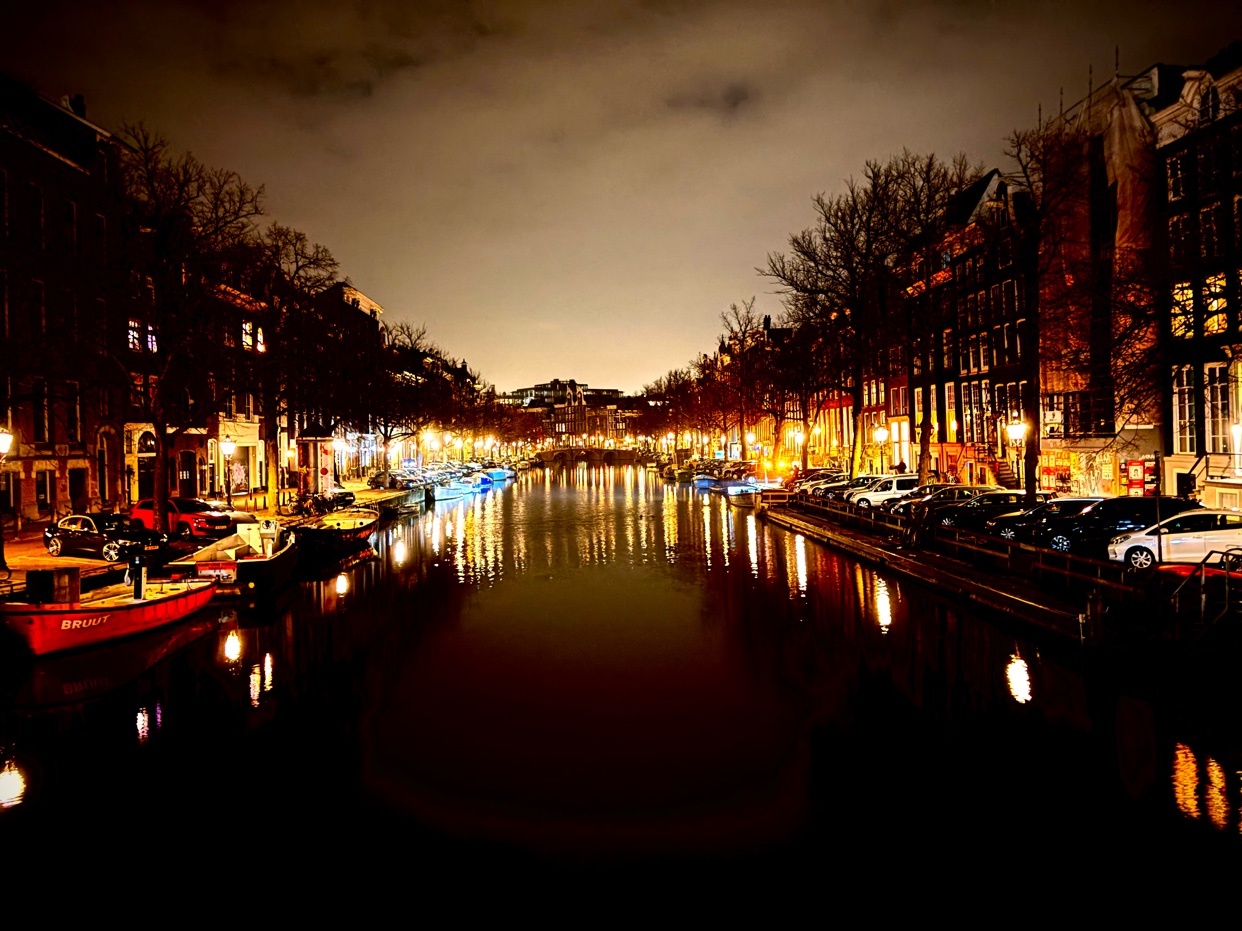 A nighttime view of an Amsterdam canal, with buildings lining the water's edge. The scene is illuminated by streetlights and reflections in the water.