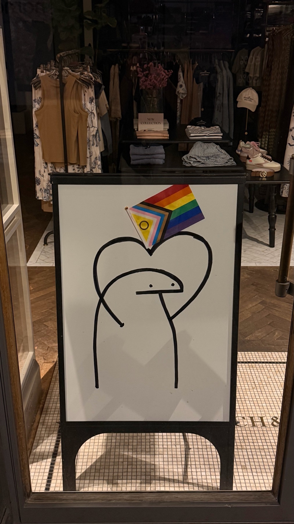 A Flork of Cows meme drawing on a whiteboard sign is displayed in front of a store. The drawing features a heart shape with a Flork character inside, topped with a pride flag.