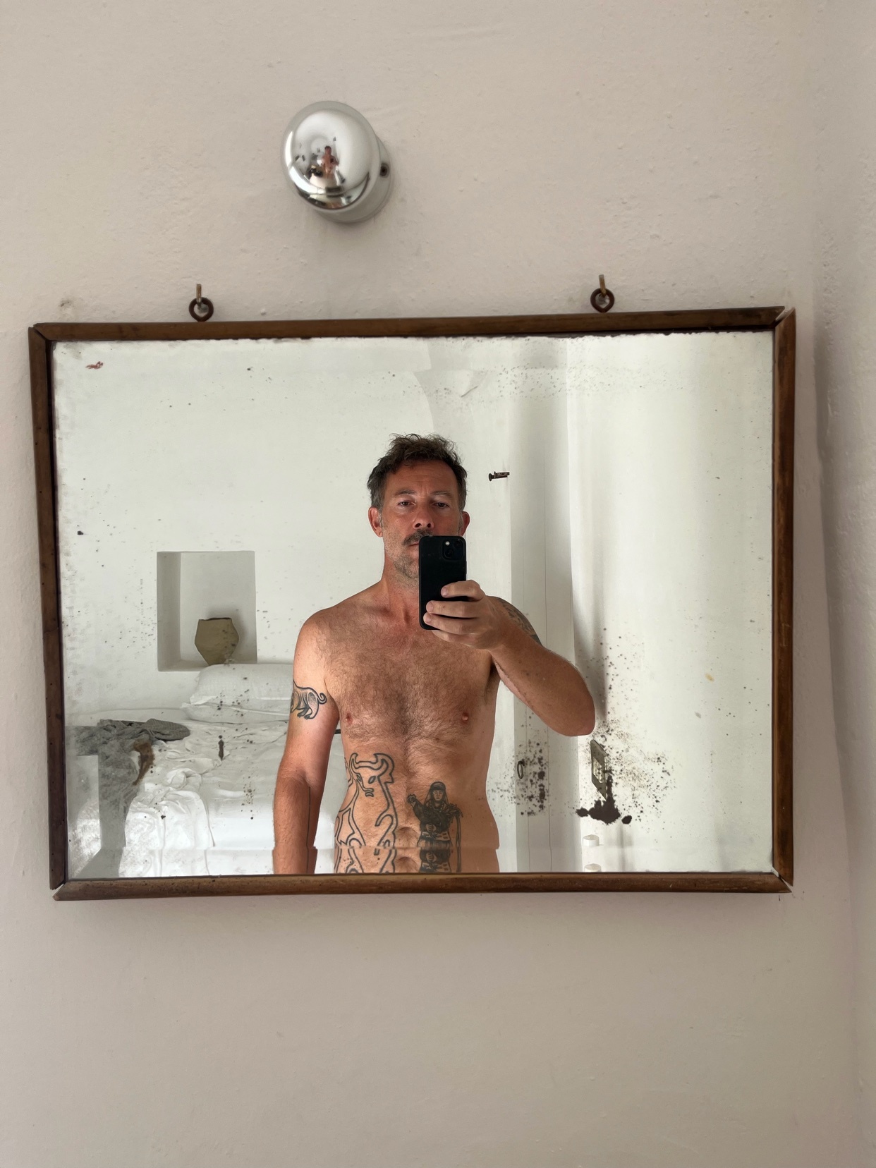 A man with tattoos takes a mirror selfie in a rustic bathroom. He is shirtless and has stubble and a mustache, with the caption 'Summer is coming'.