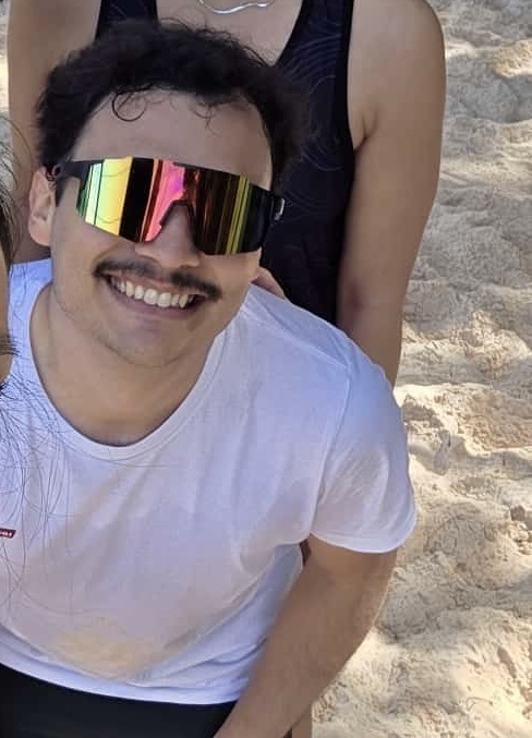 A man with a mustache and large, colorful sunglasses smiles at the camera while wearing a white t-shirt and black shorts. He appears to be at a beach with sand visible in the background.