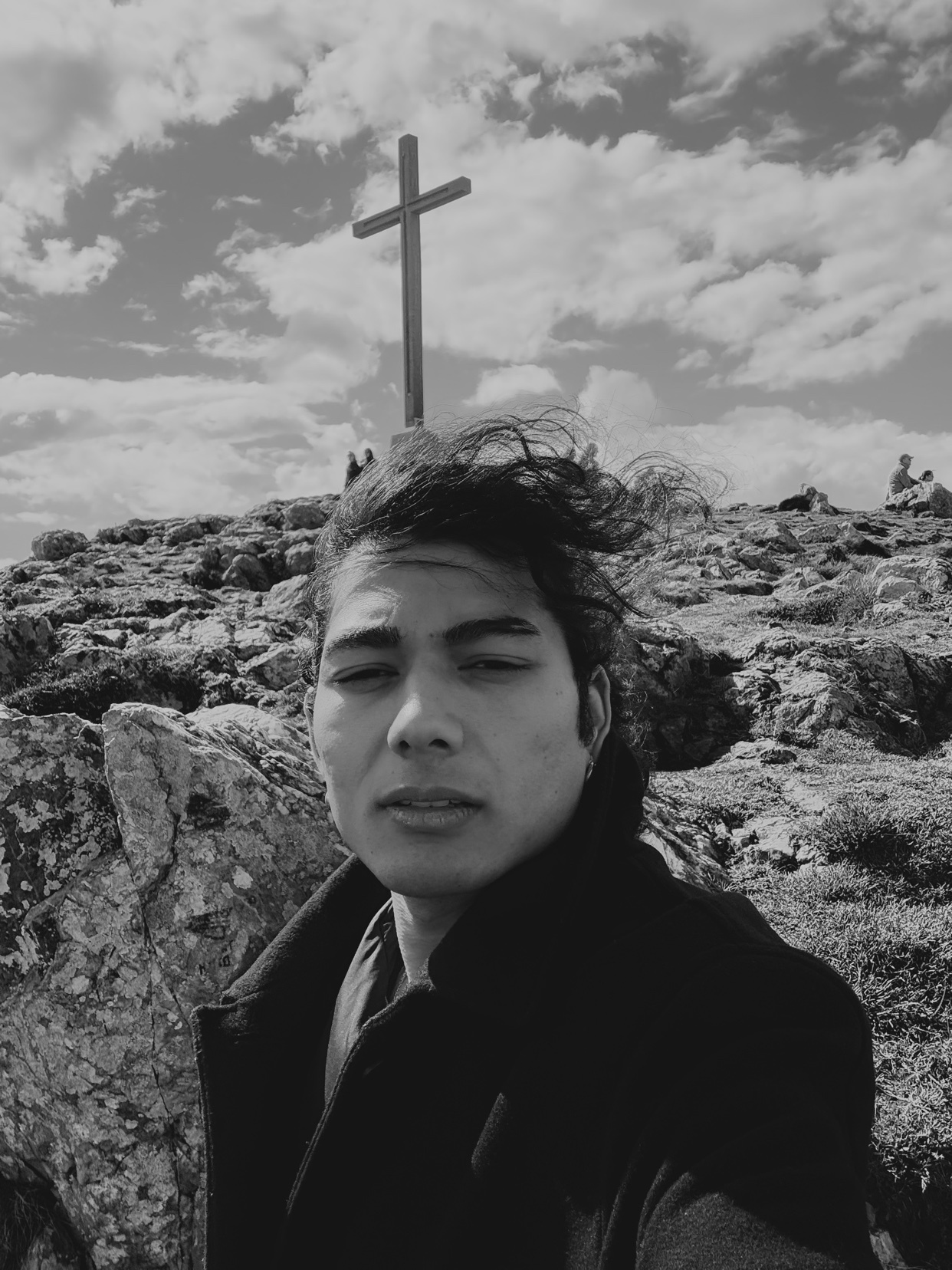 A man takes a selfie on a rocky mountain with a large cross in the background under a cloudy sky.