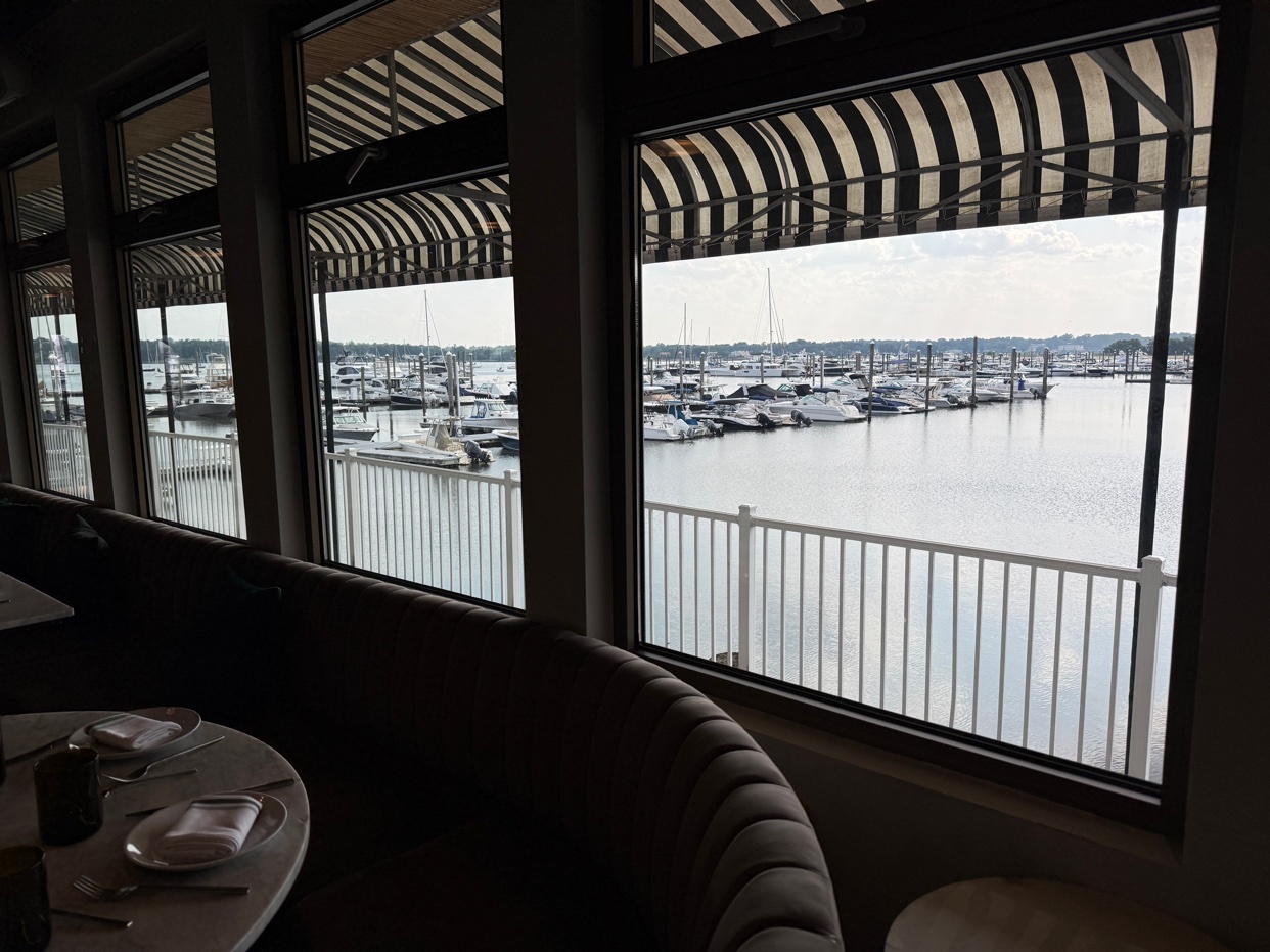 A restaurant interior with a view of a marina. The windows are framed with black and white awnings.