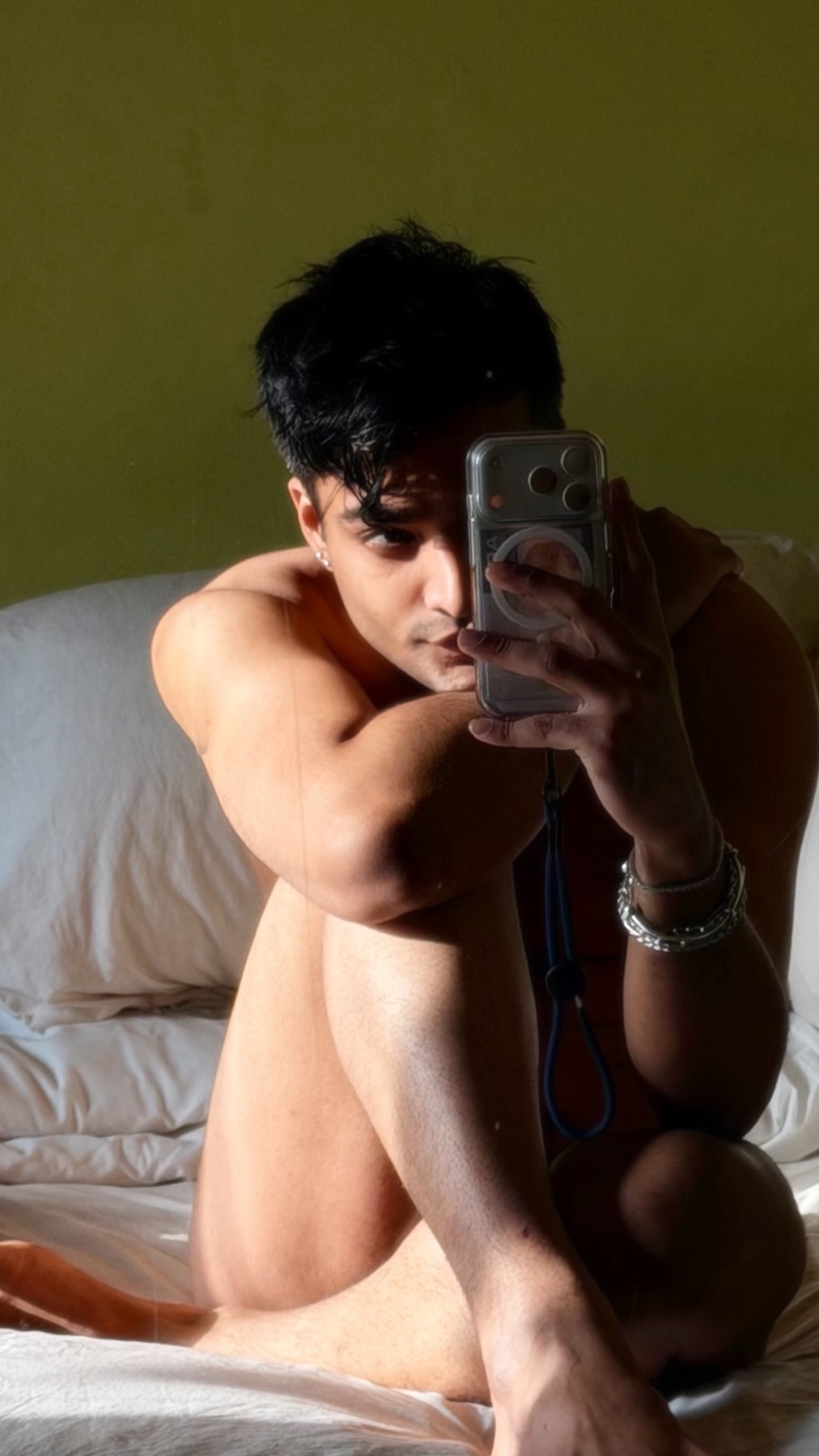 A man takes a mirror selfie while sitting on a bed. He is shirtless and looking at the camera.