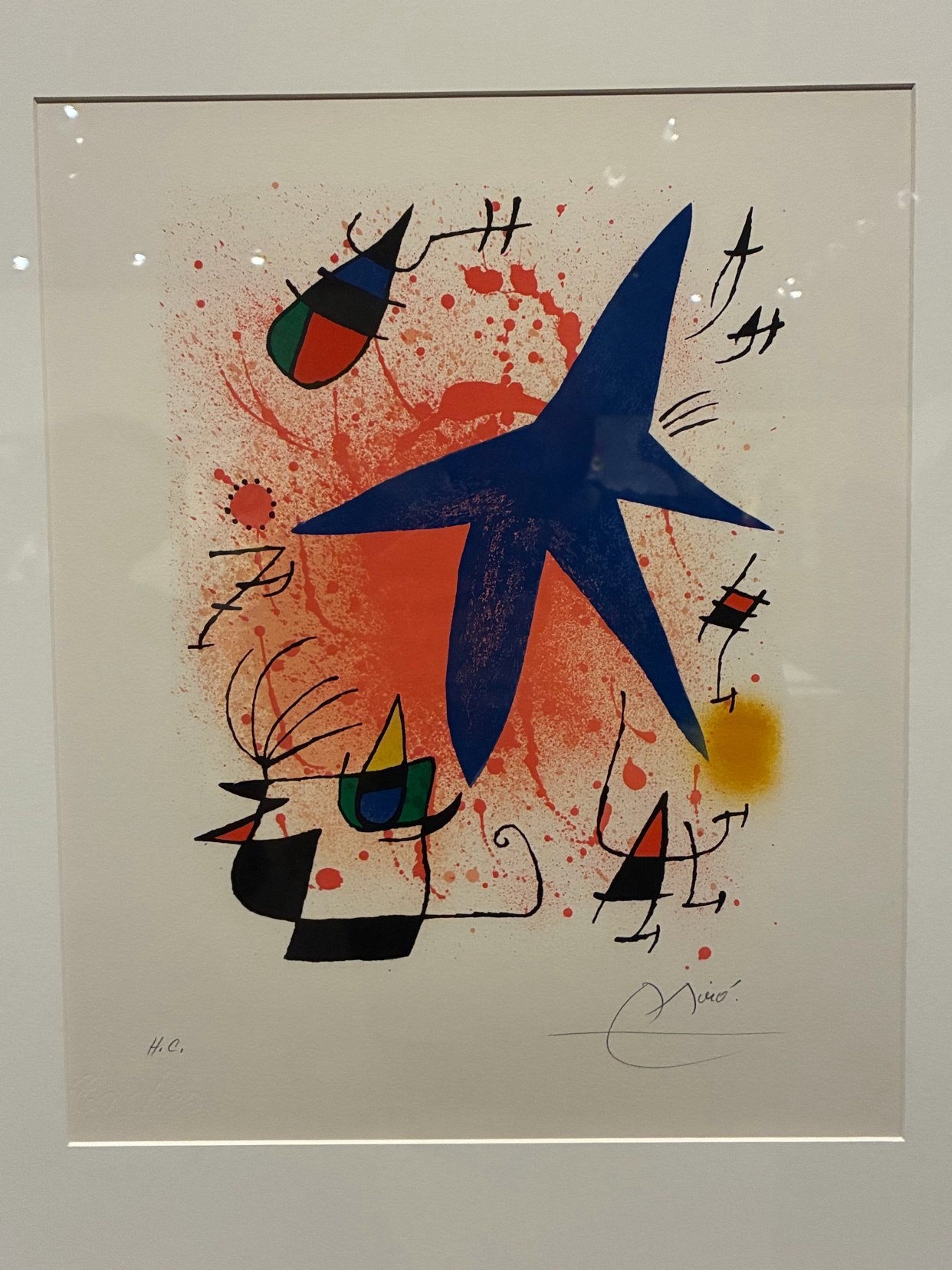 A vibrant abstract lithograph by Joan Miró features a large blue starburst at the center, surrounded by playful, biomorphic shapes and energetic red splatters. The artwork is signed by the artist in the lower right corner.