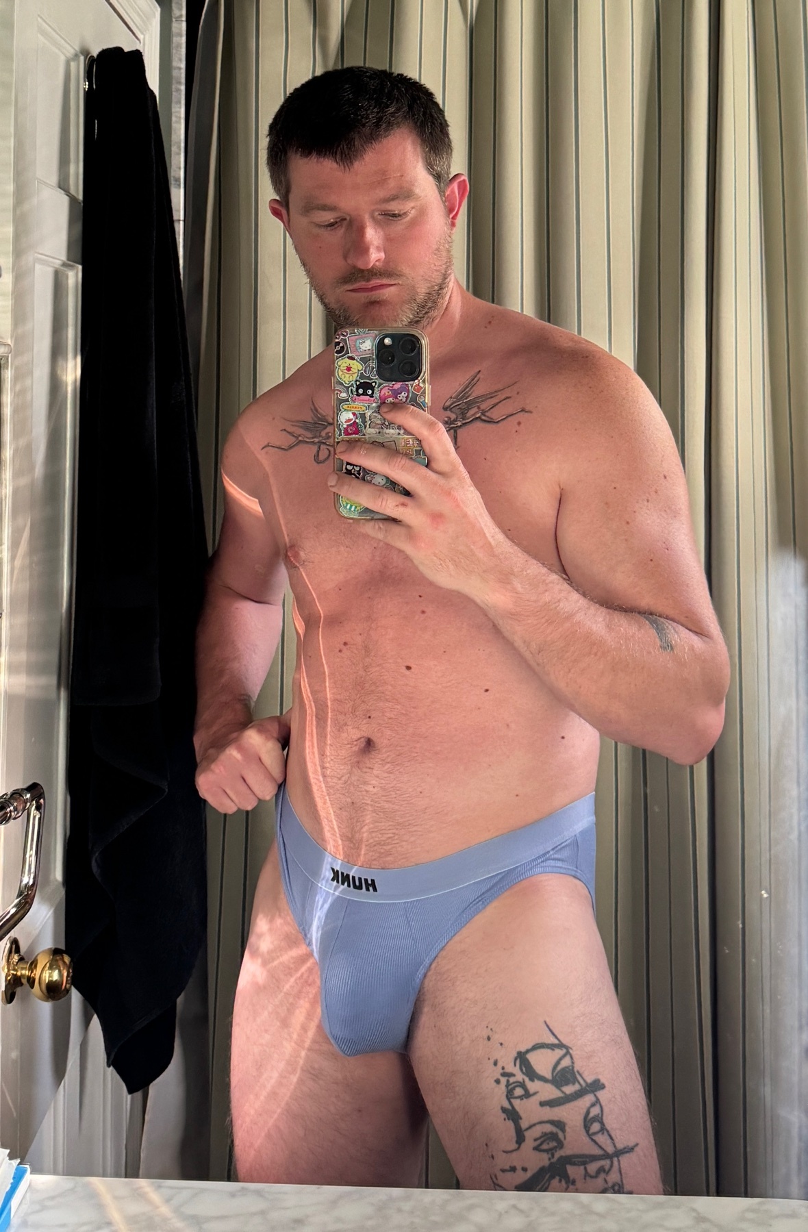 A man in blue briefs takes a mirror selfie in a bathroom. He has tattoos on his chest and thigh.