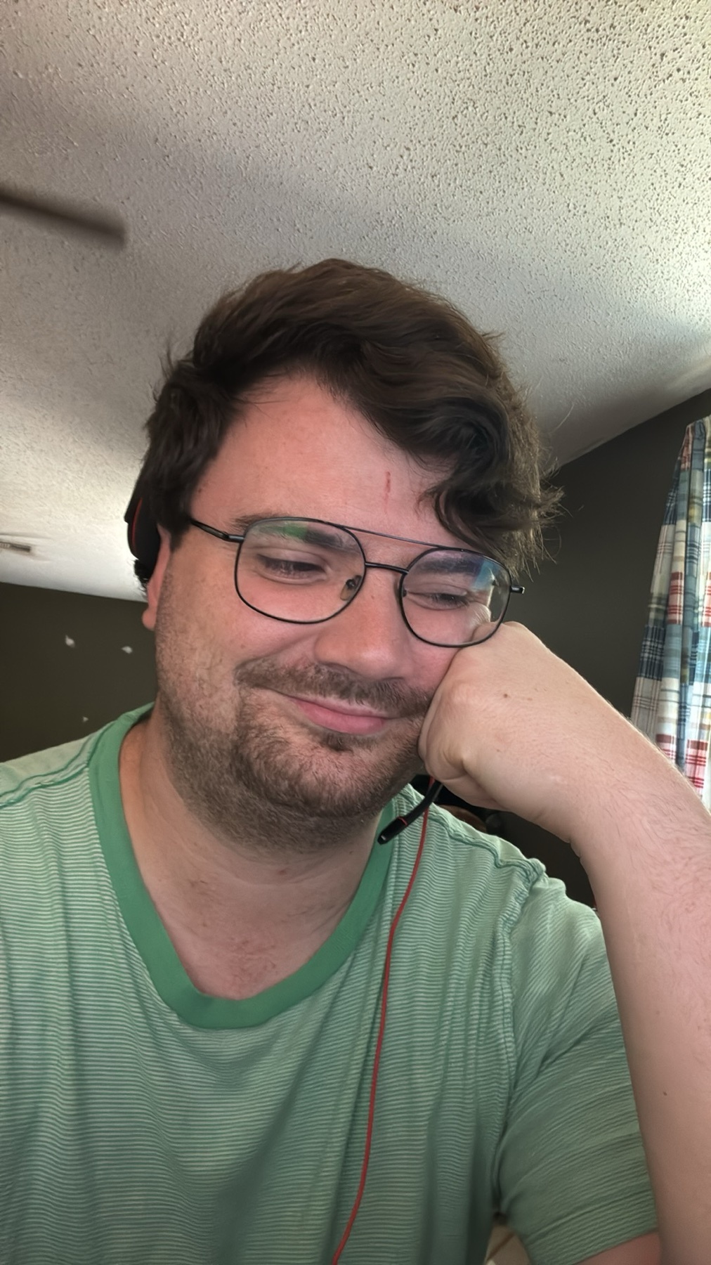 A man with short, brown hair and glasses rests his head on his hand, smiling with his eyes closed. He is wearing a green t-shirt.