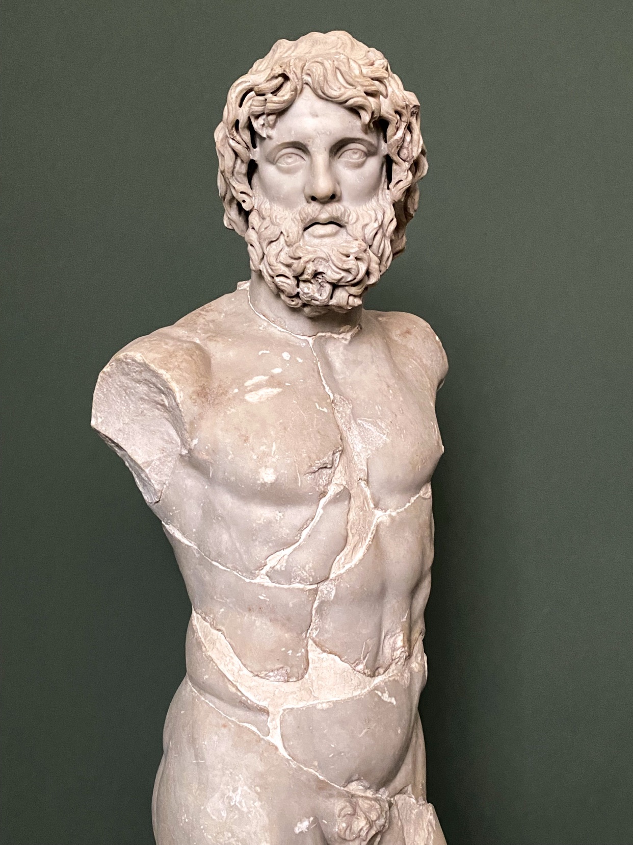 A marble sculpture of a muscular man with a beard and curly hair stands against a dark green background. The sculpture is fragmented, with visible cracks across the torso and lower body.