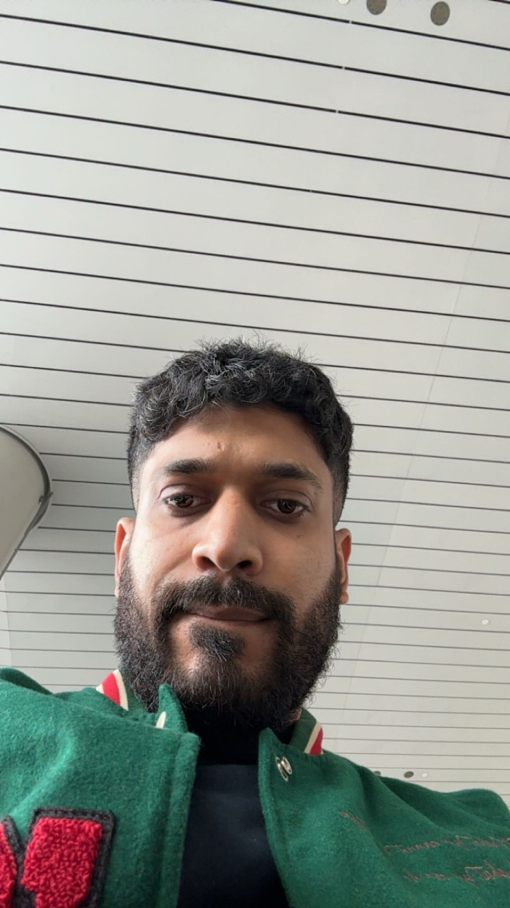 A man with a beard and curly dark hair takes a selfie in front of a white wall with horizontal black lines.