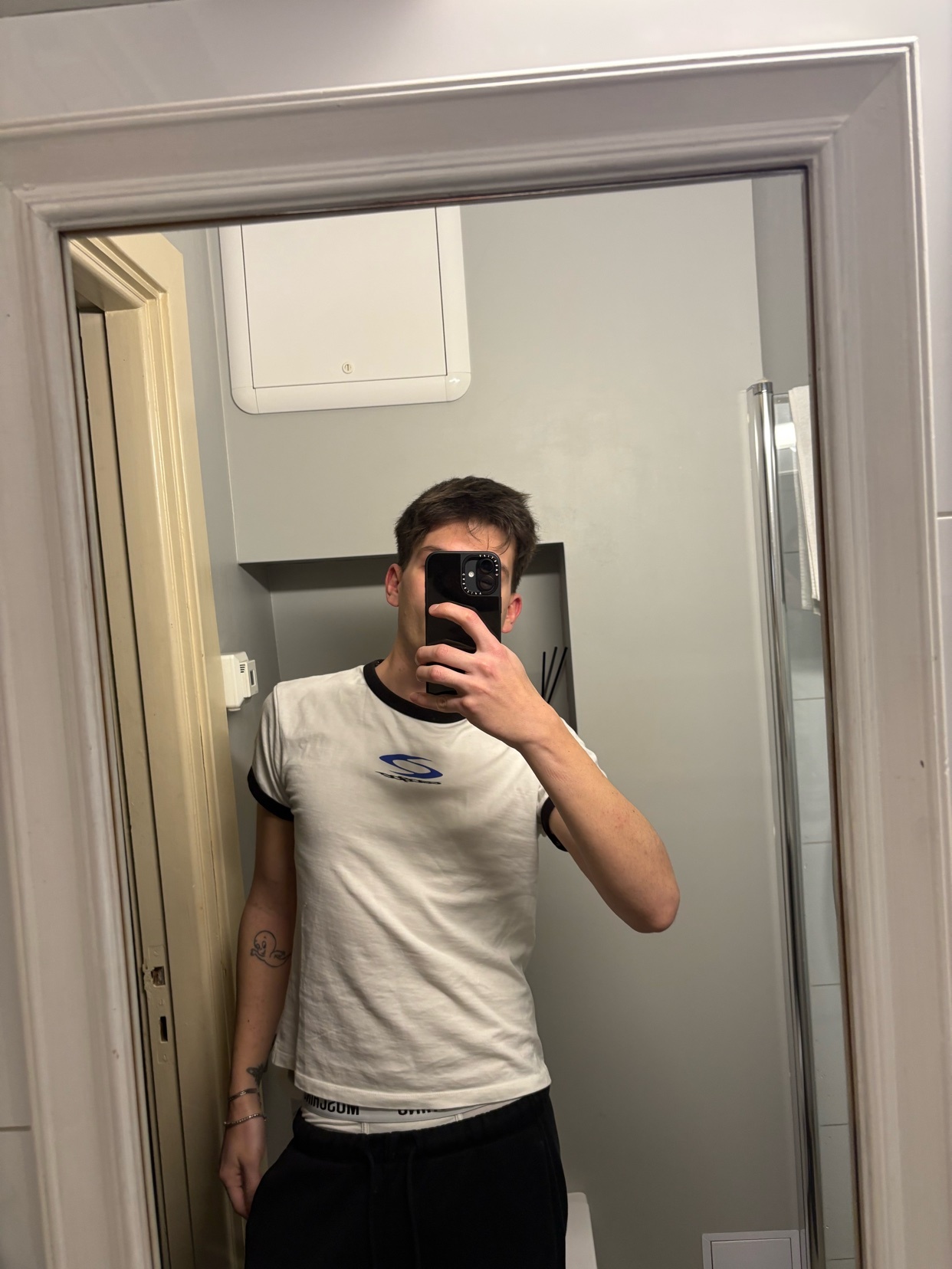 A man takes a selfie in a bathroom mirror, wearing a white t-shirt with black trim and dark pants. He has a tattoo on his left arm.