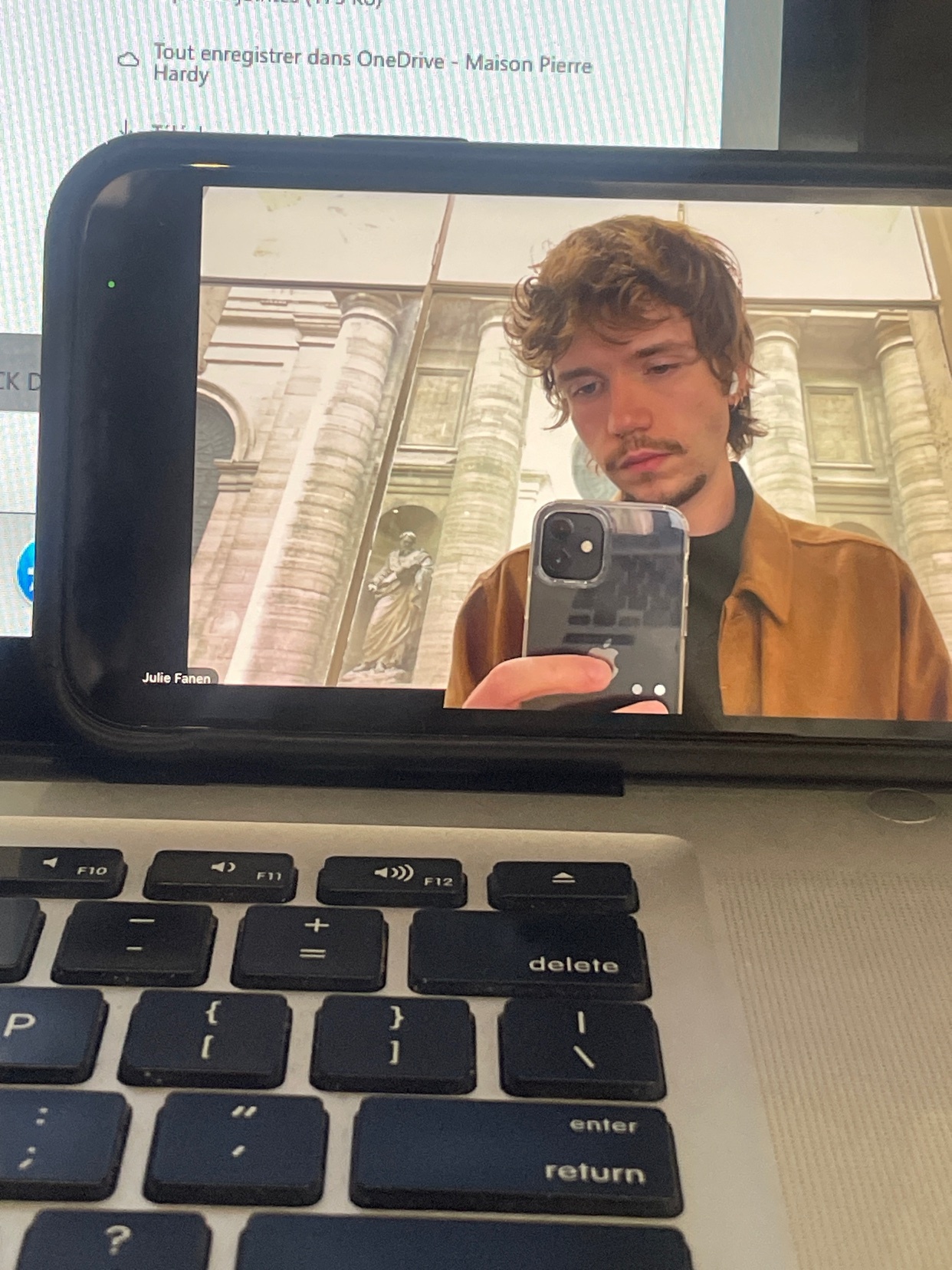 A man takes a selfie in front of a building with large columns. The photo is taken with a phone, and the man is looking at the camera.