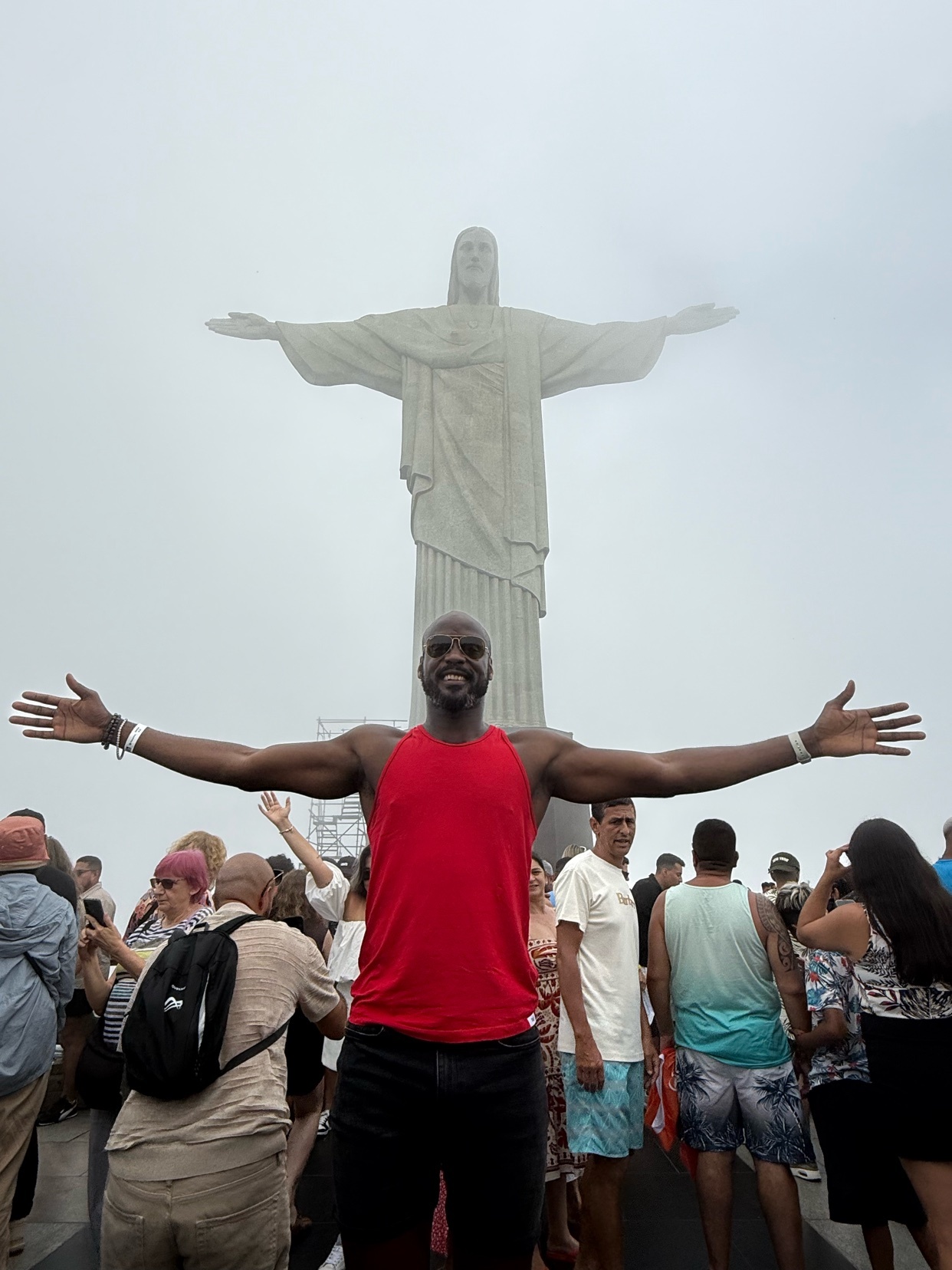 A man poses with arms outstretched in front of the Christ the Redeemer statue in Rio de Janeiro, Brazil. The man is shirtless and smiling.