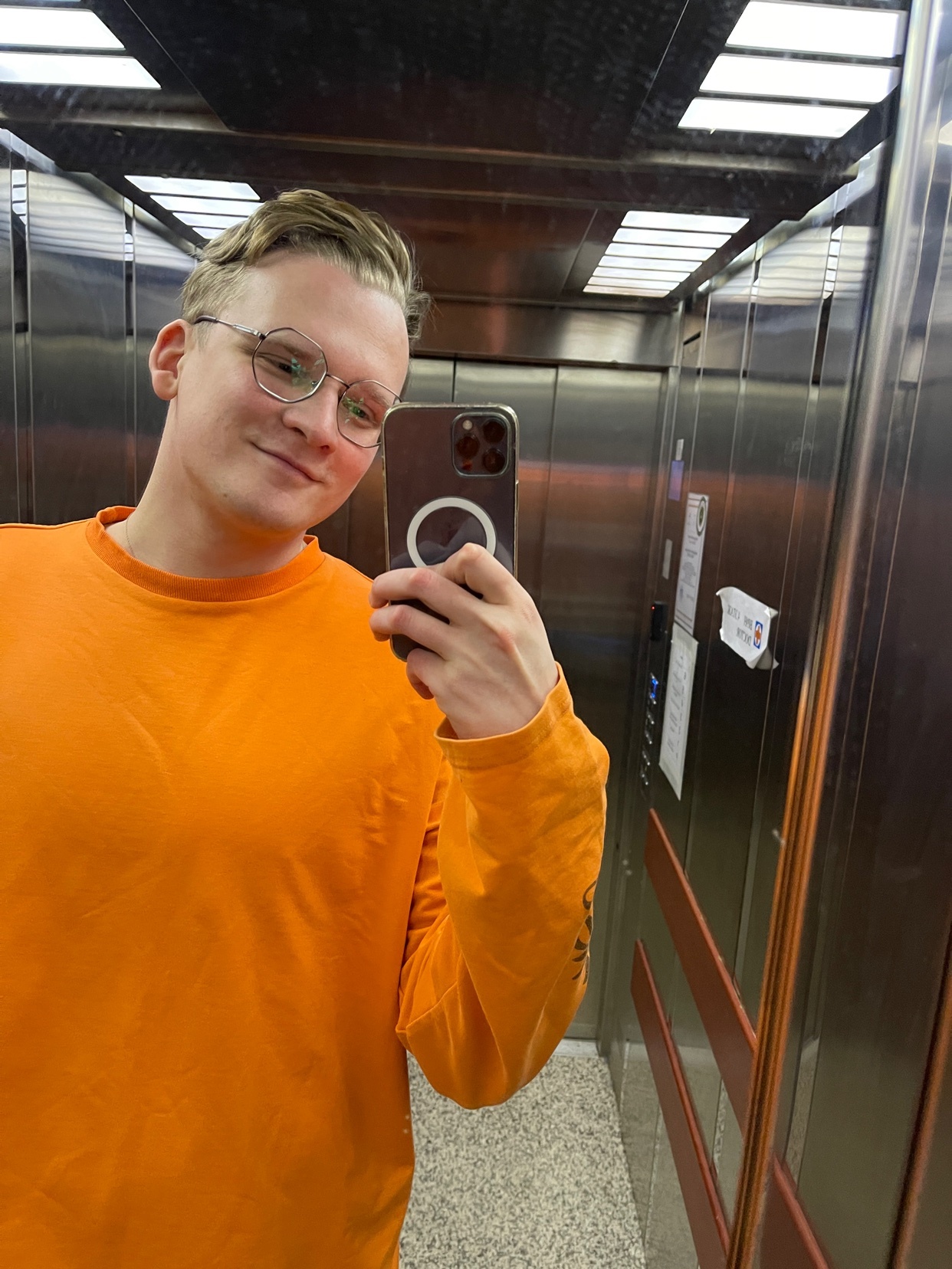A man takes a selfie in an elevator, wearing glasses and an orange long-sleeved shirt. The mirror reflects the interior of the elevator.