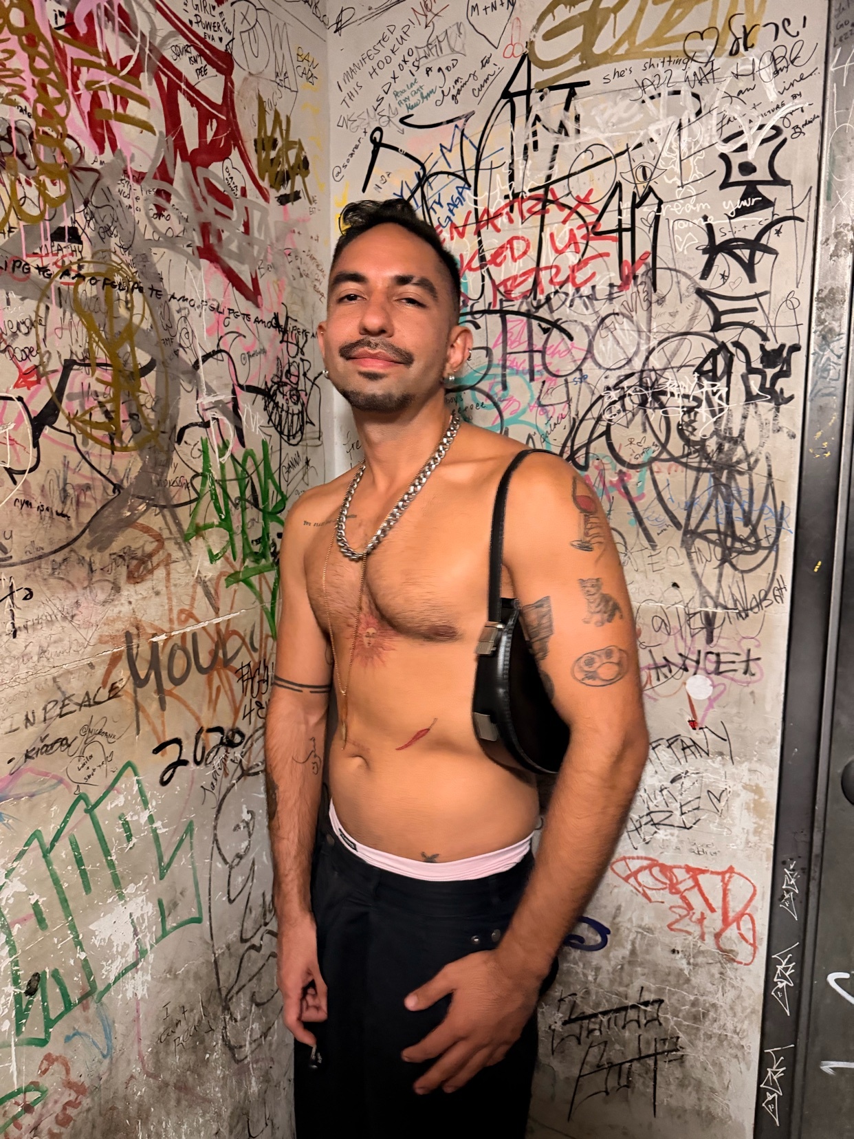 A shirtless man with tattoos and a moustache poses in front of a graffiti-covered wall. He is wearing a silver chain and a black shoulder bag.