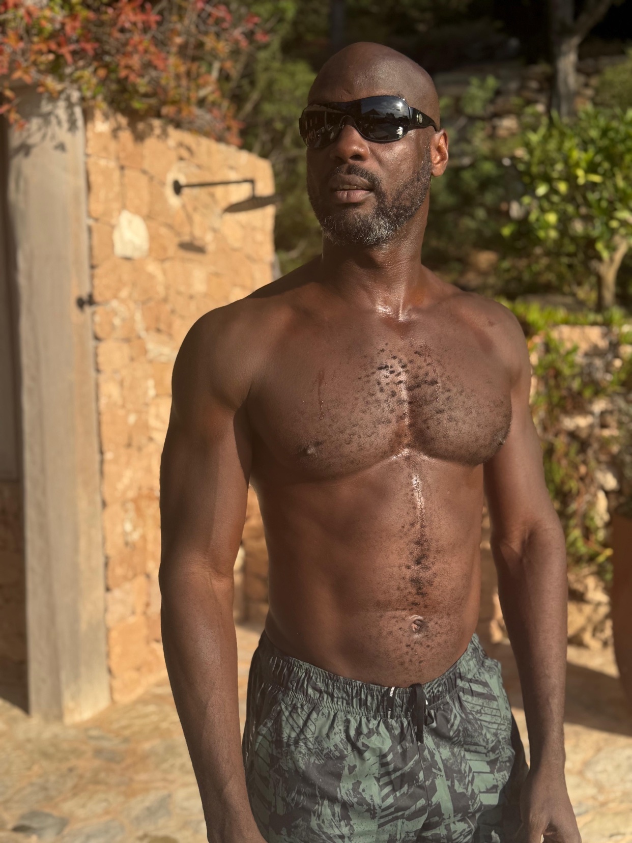 A shirtless man with a muscular physique poses outdoors. He is wearing sunglasses and swim trunks.
