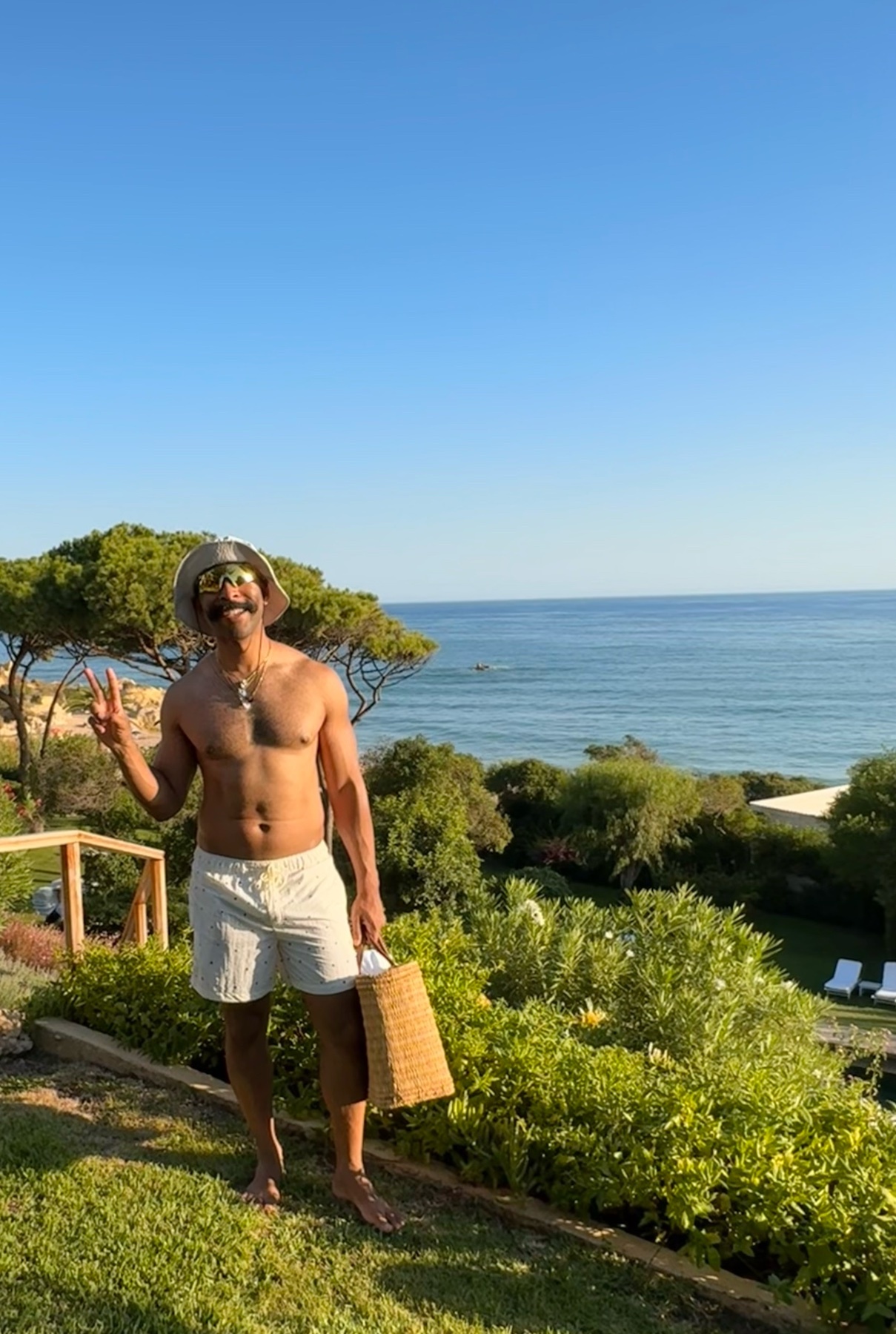A man in a hat and sunglasses poses for a photo on a grassy hill overlooking the ocean. He is wearing striped shorts and holding a woven bag, giving a peace sign with his left hand.