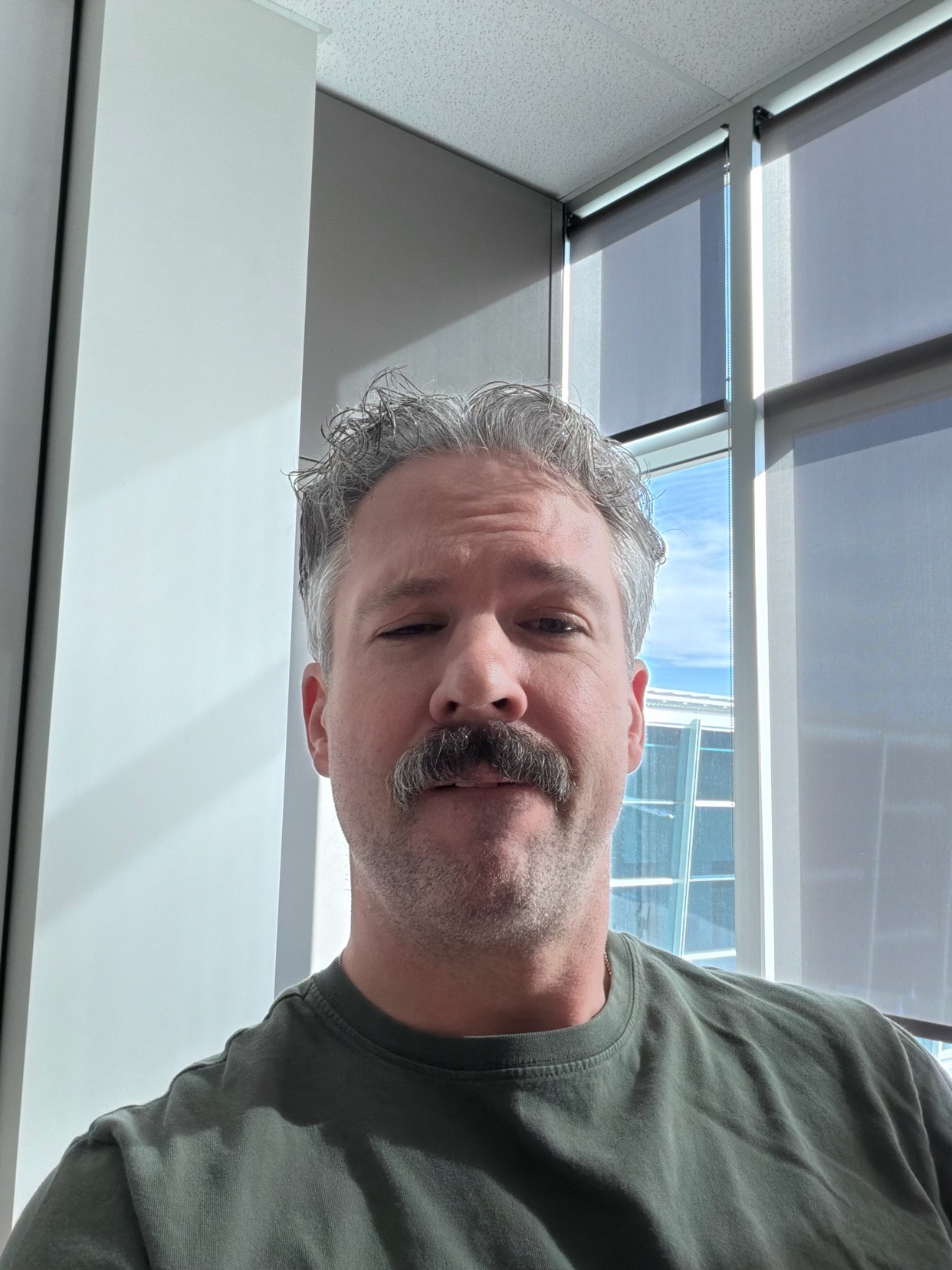 A man with a prominent mustache and grey hair takes a selfie indoors, with blinds covering a window in the background.