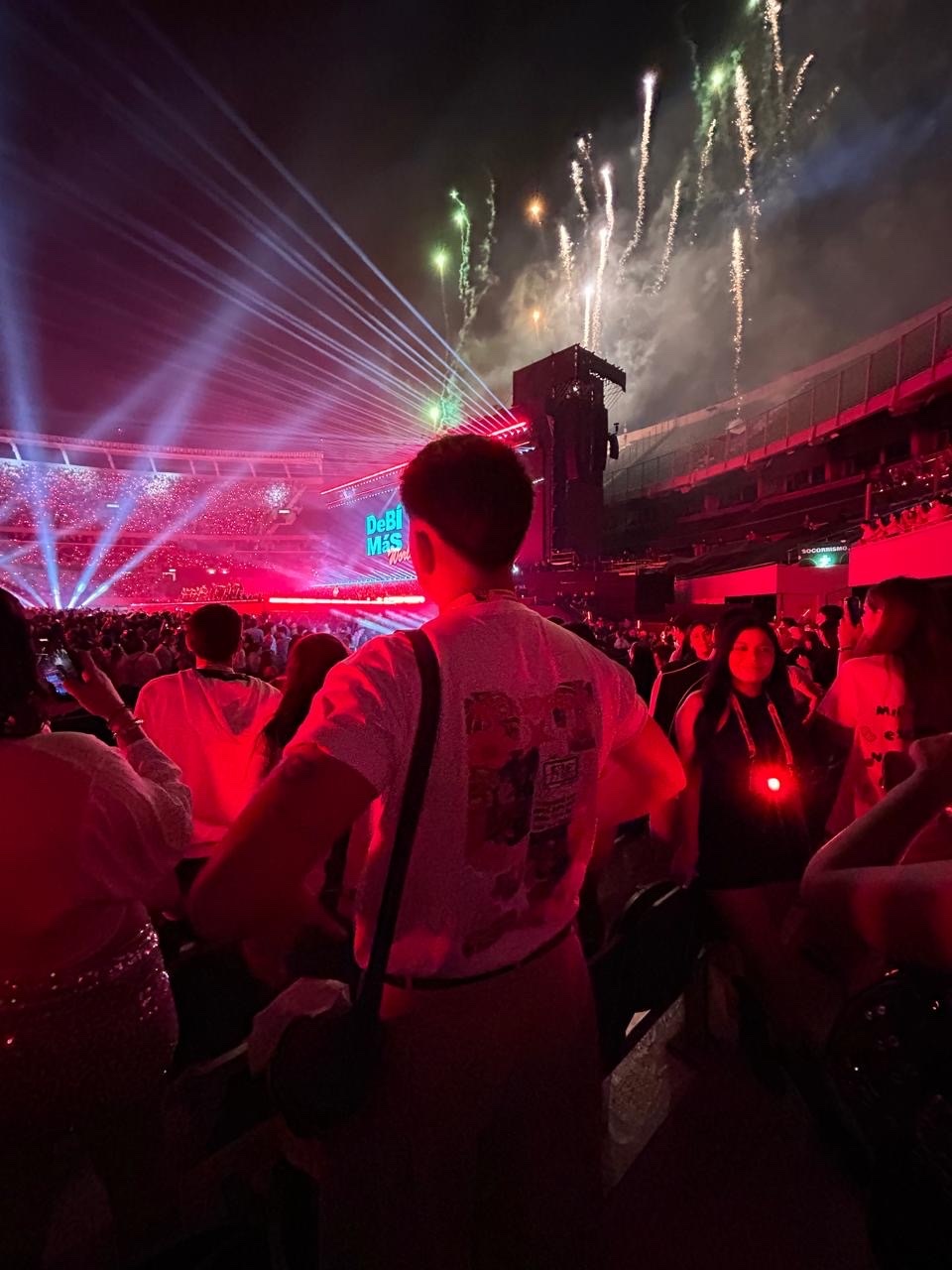 A person watches fireworks at a Bad Bunny concert. The photo is taken from behind the person, showing the crowd and stage.