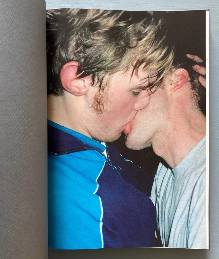 A close-up photograph captures two men kissing passionately. The image is part of a book, with the left page showing a textured grey background.