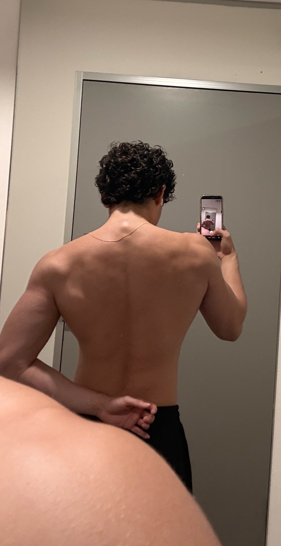 A man with curly hair takes a mirror selfie showing his back. He is shirtless and appears to be in a gym or locker room.