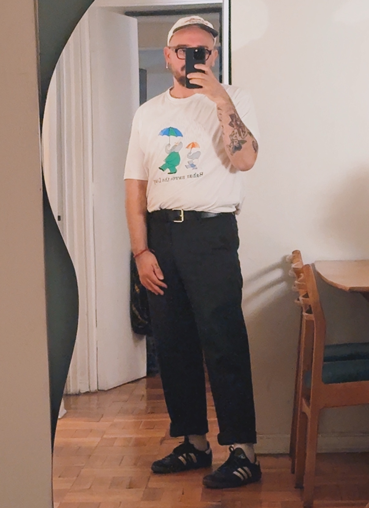 A man takes a mirror selfie, showcasing his outfit. He wears a graphic t-shirt, black pants, and a cap.