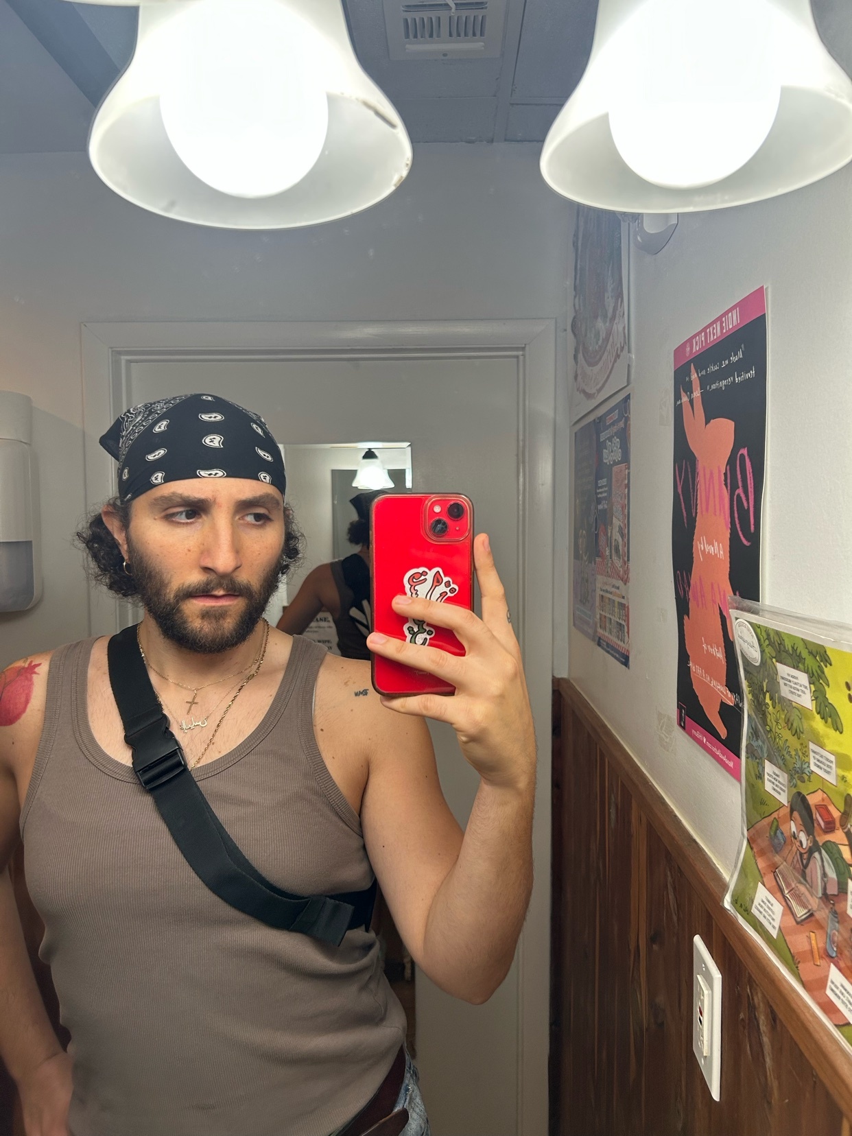 A man with curly hair and a bandana tied around his head takes a selfie in a bathroom mirror. He is wearing a brown tank top and a cross necklace.
