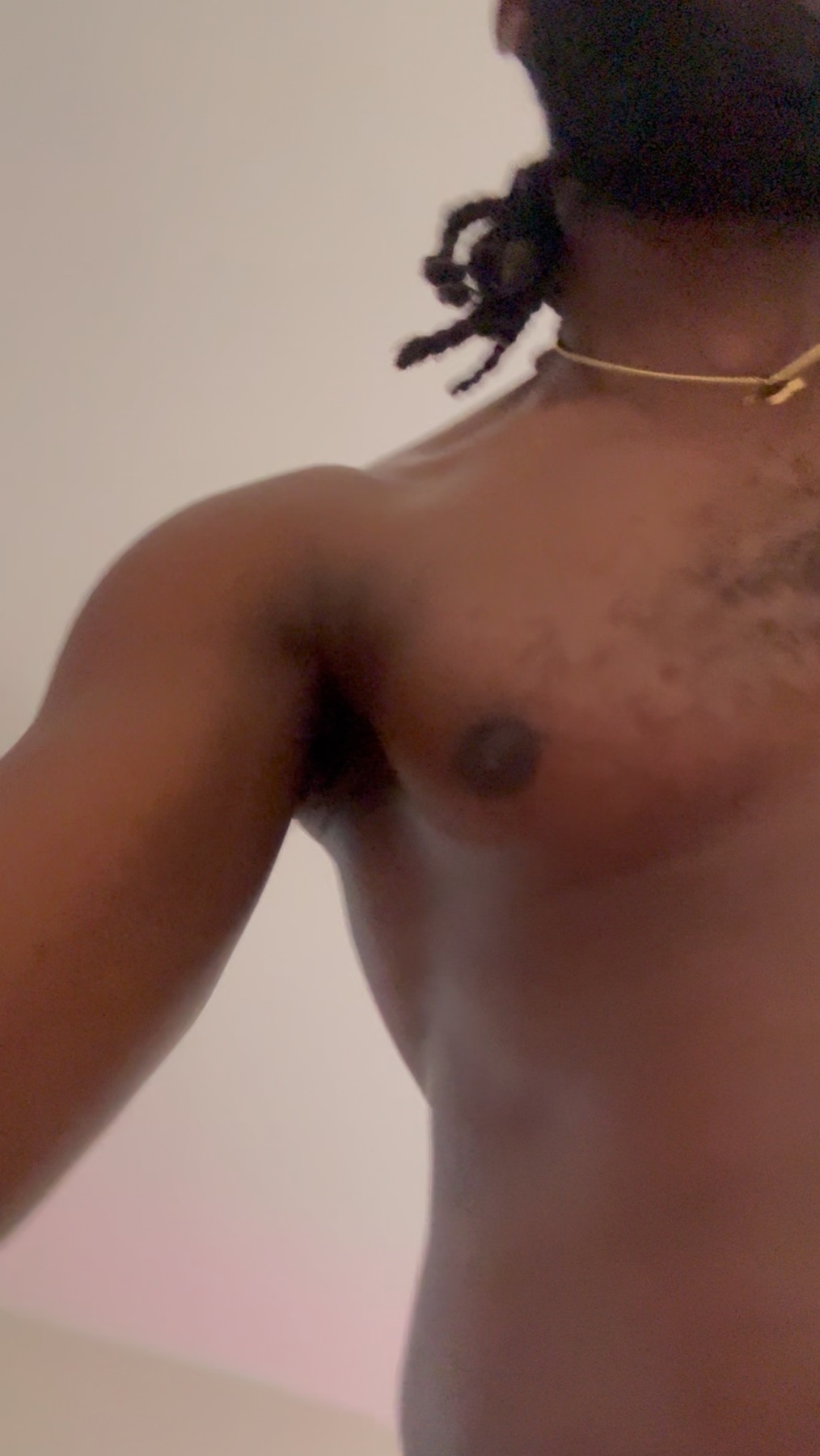 A close-up, low-angle shot of a shirtless Black man with dreadlocks and a necklace. He has visible chest hair and appears to be flexing or posing.