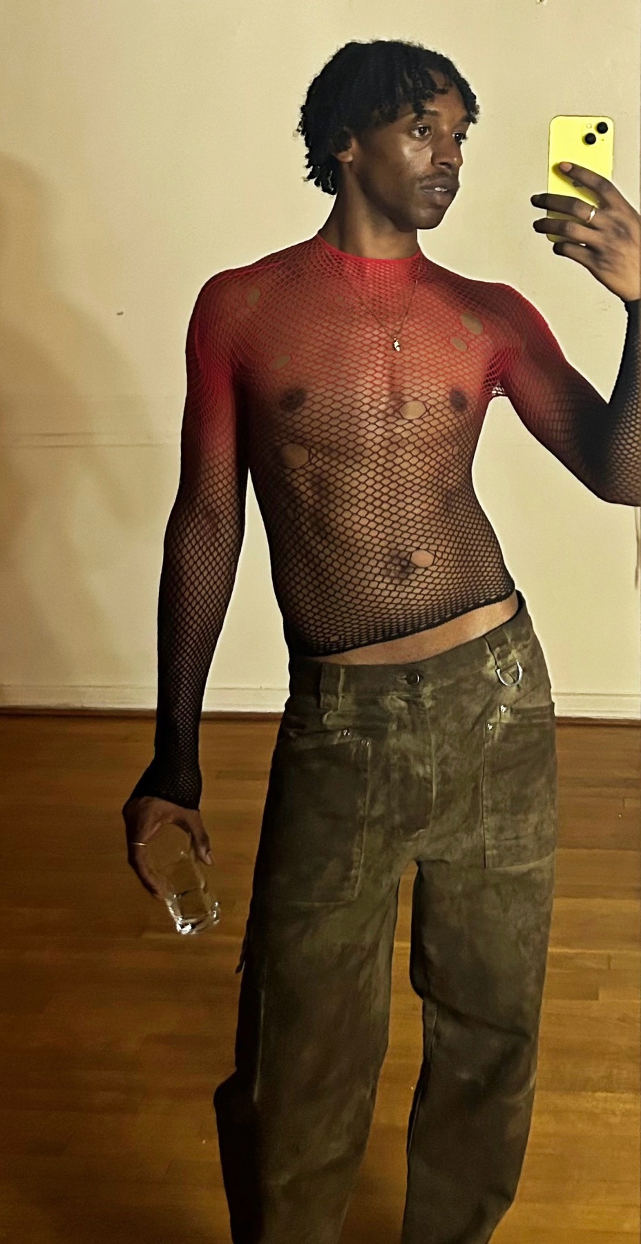 A man takes a mirror selfie, showcasing a bold fashion statement. He is shirtless, wearing a fishnet top and stylish pants.