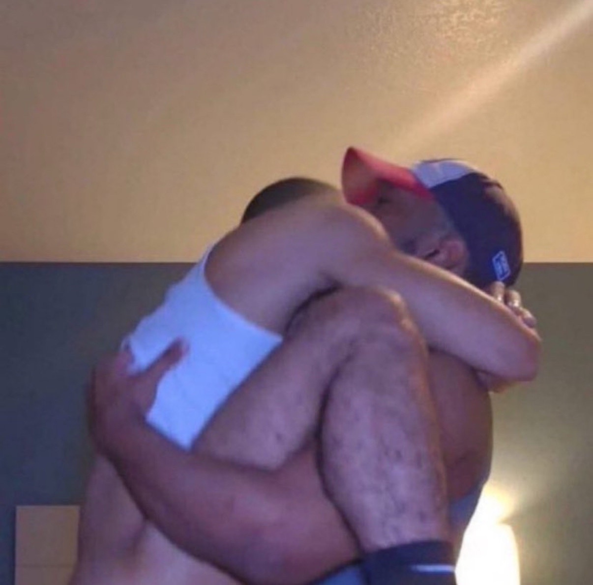 A couple embraces in a bedroom. One man is shirtless, and the other is wearing a tank top and a cap.
