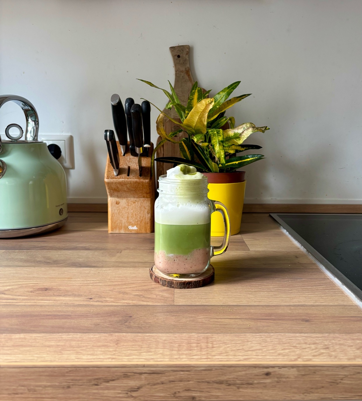 A layered strawberry banana matcha latte sits on a wooden coaster on a kitchen counter. The drink is in a mason jar with a handle and topped with whipped cream. A knife block, a plant in a yellow pot, and a kettle are visible in the background.