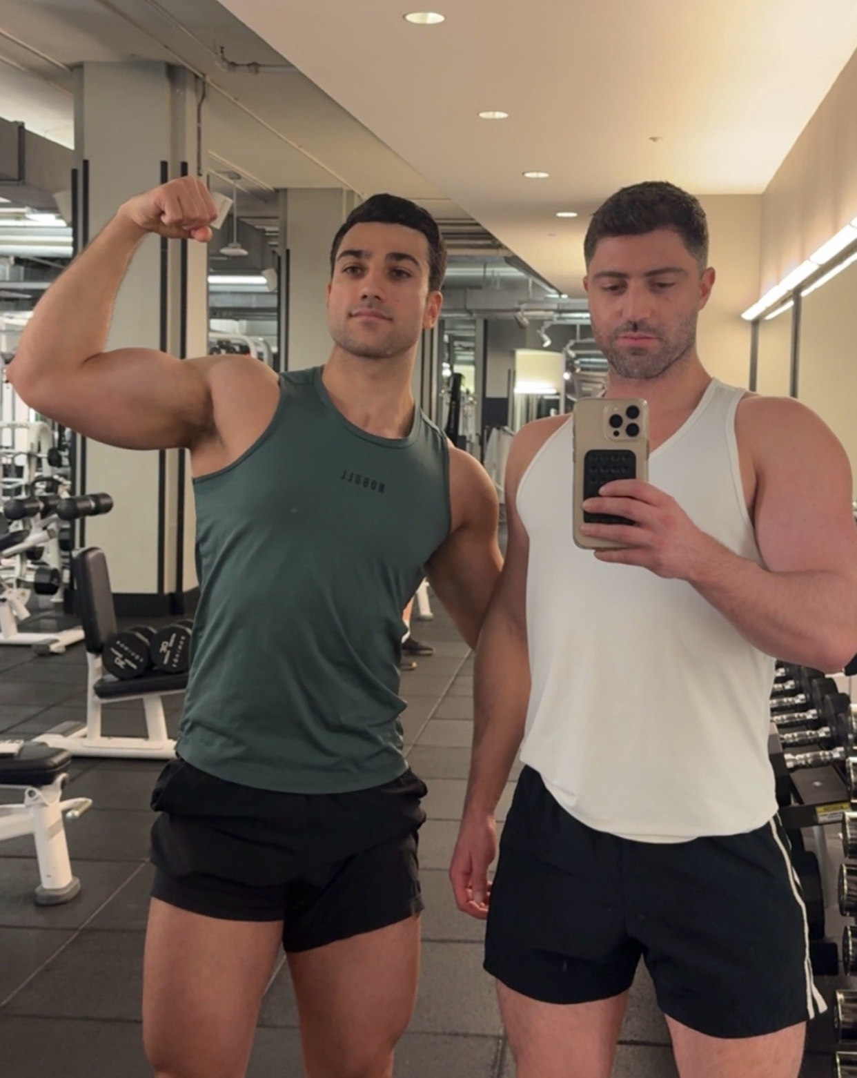 Two muscular men pose for a selfie in a gym. One man flexes his bicep while wearing a dark green tank top and black shorts. The other man, wearing a white tank top and black shorts, holds the phone to take the picture.