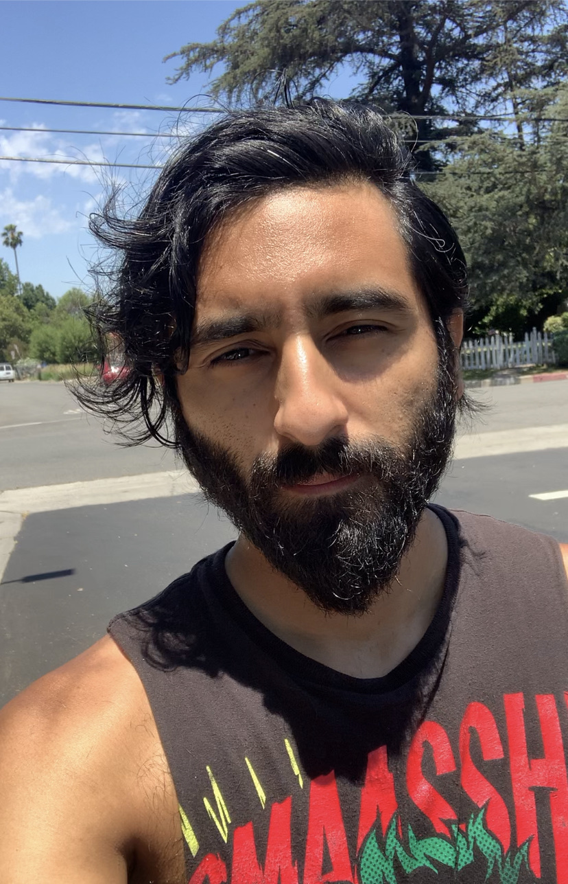 A man with a beard and dark, wavy hair takes a selfie outdoors on a sunny day. He is wearing a dark, sleeveless t-shirt with red and green lettering.