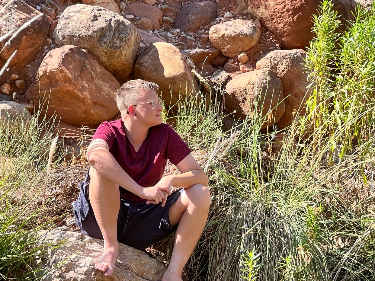 A young man with glasses sits on a rock at the bottom of the Grand Canyon, looking off to the side. He is wearing a maroon t-shirt and dark shorts, with lush green plants and large rocks surrounding him.